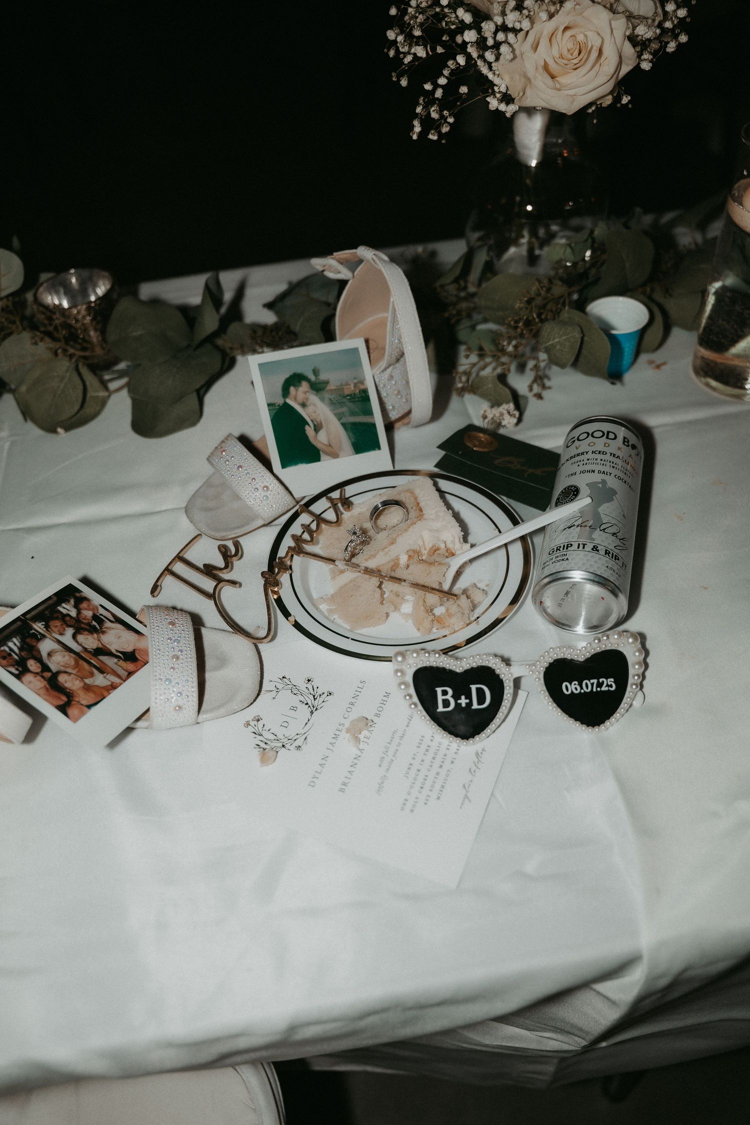 Wedding reception table details with decorative hearts, photographs, a white rose centerpiece and Jack Daniels bottle on white tablecloth.