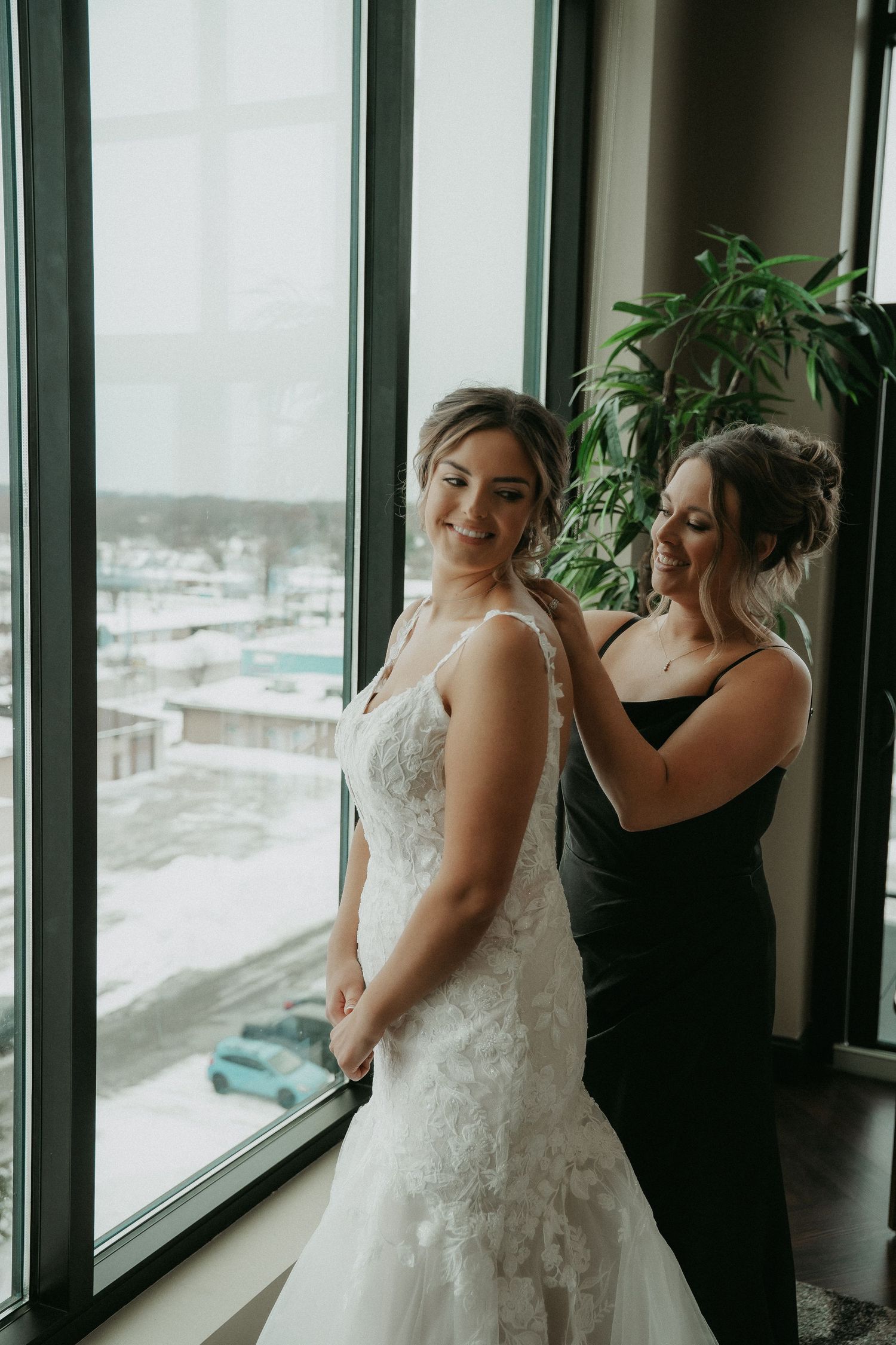 A bride in a white lace wedding dress gets ready by a large window with winter scenery outside.