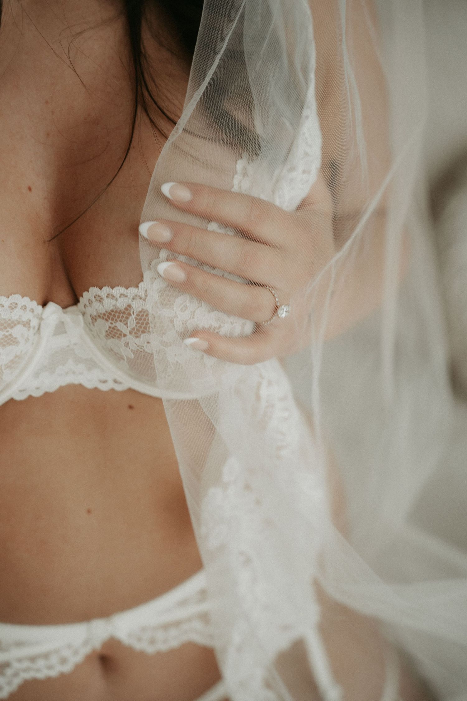 Bride in white lace lingerie holding a sheer veil, showcasing an elegant engagement ring on her hand.