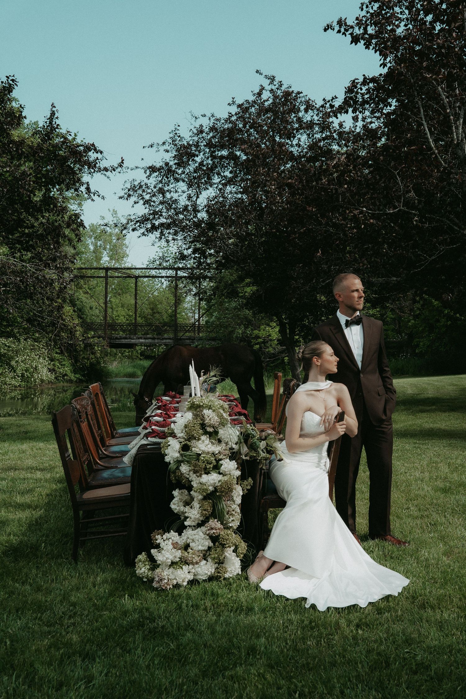 Bride in white gown seated at outdoor floral table with groom in tuxedo standing beside her on lush green lawn.