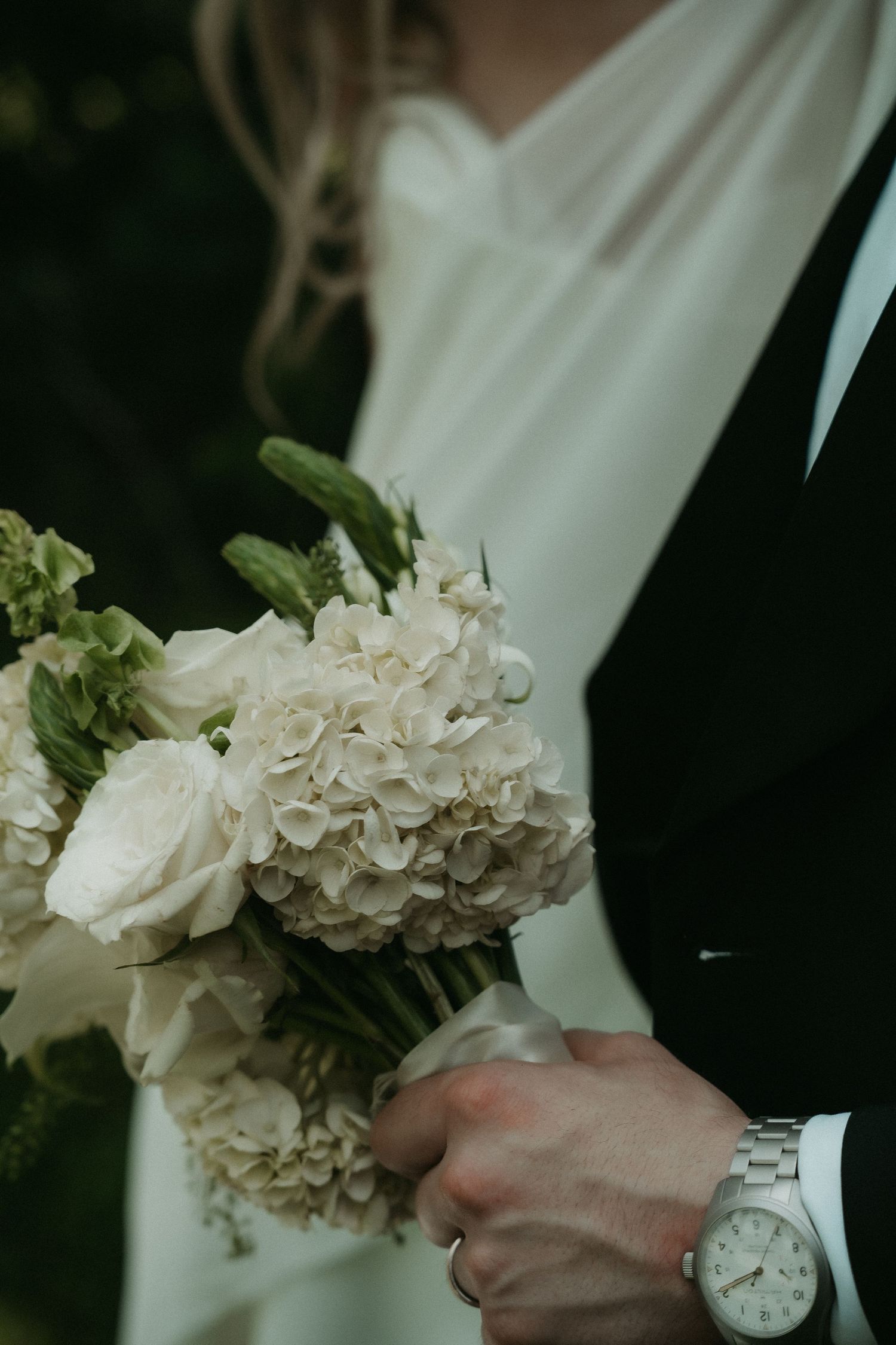 Bride and groom holding a white hydrangea and rose wedding bouquet, groom wearing a silver watch.