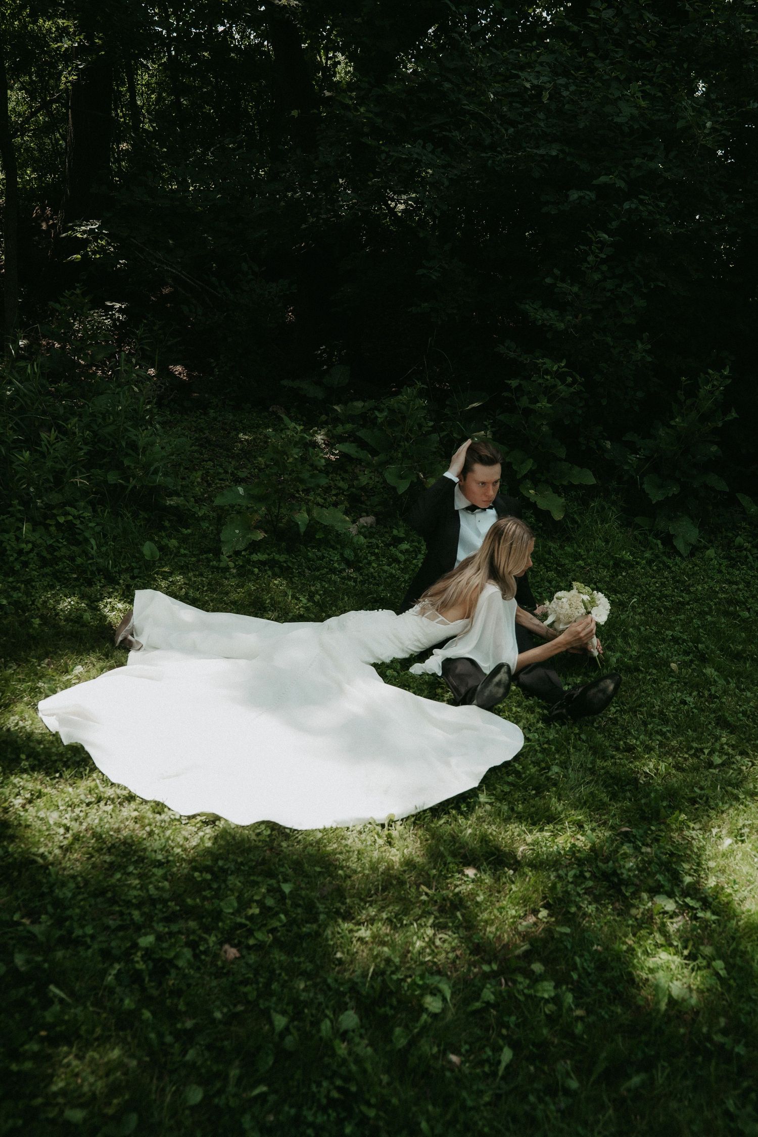 Bride and groom sitting on lush green grass in a forest, bride in flowing white gown holding a bouquet.