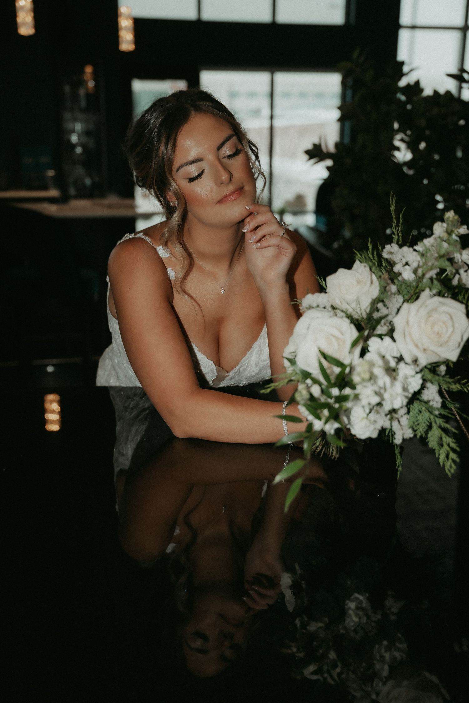 Romantic bridal portrait with white roses and greenery bouquet reflecting in dark table at moody indoor venue.