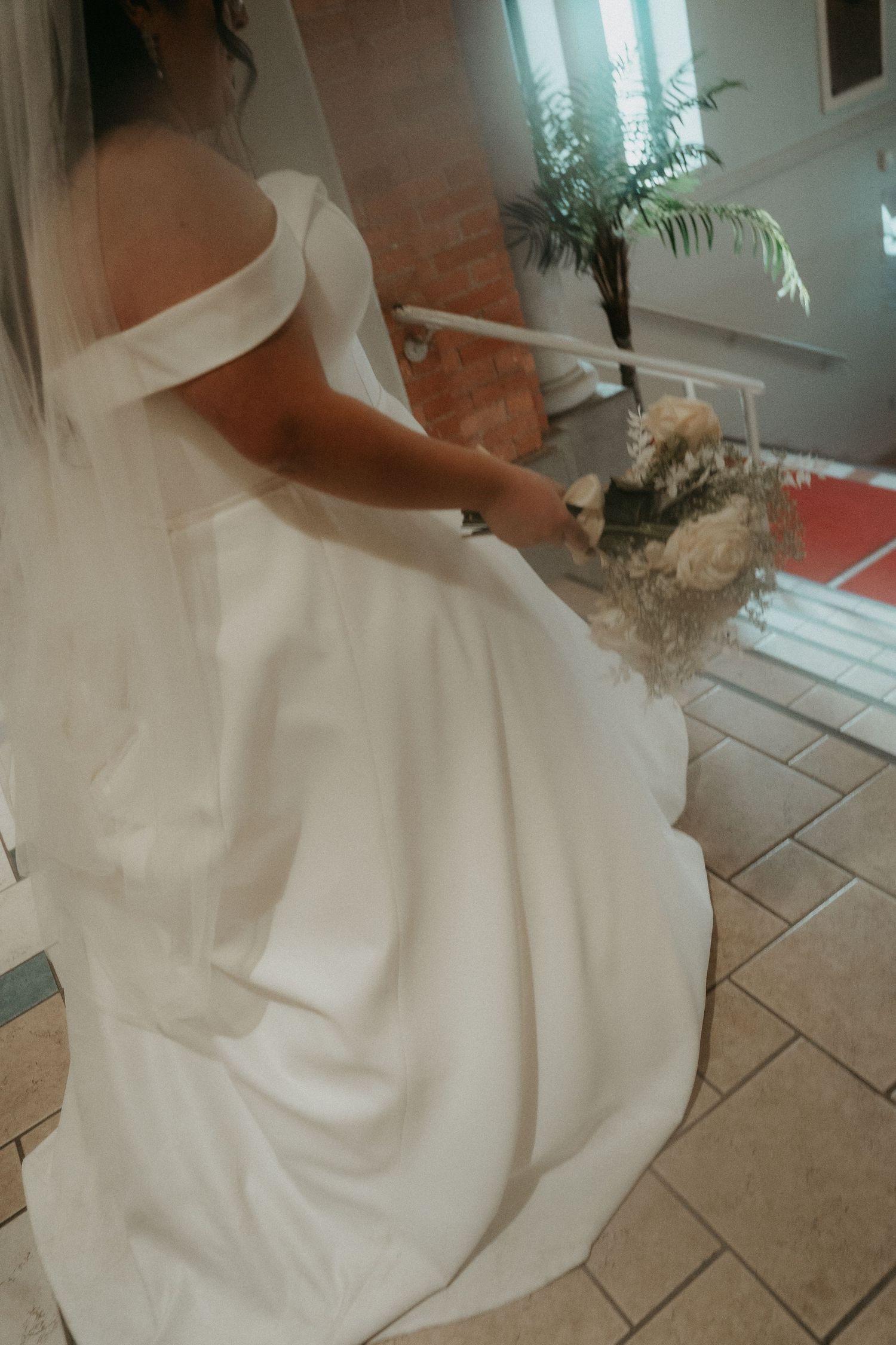 A bride in an elegant white off-shoulder wedding gown sits on tiled floor holding a bouquet.