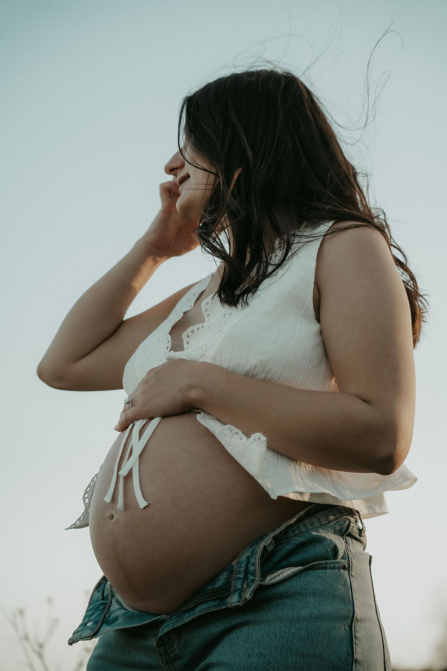 Pregnant woman in white crop top and jeans cradling her bare baby bump outdoors against a clear sky.