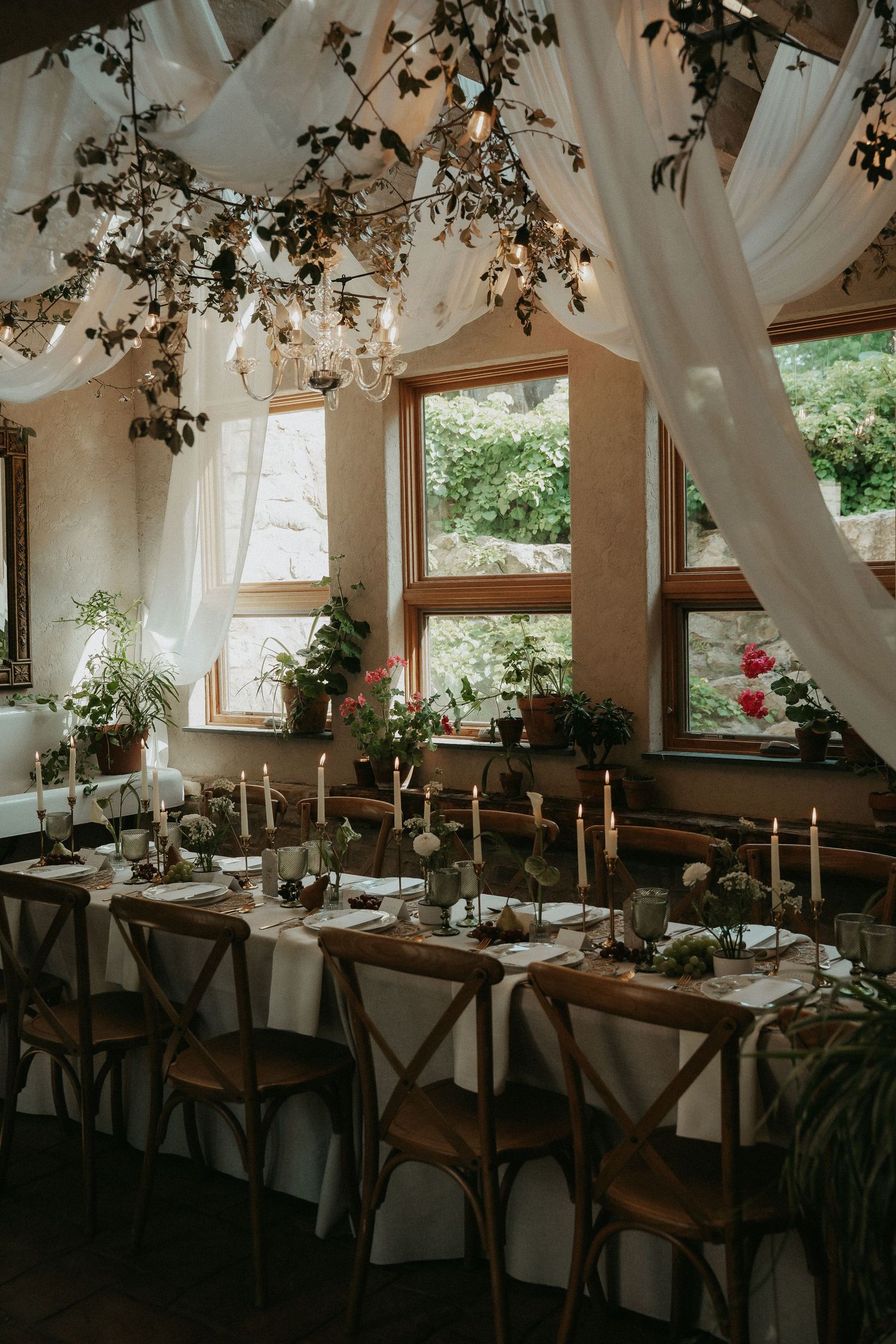 Elegant wedding reception with wooden cross-back chairs, candlelit long table, white draping, and lush greenery decor.