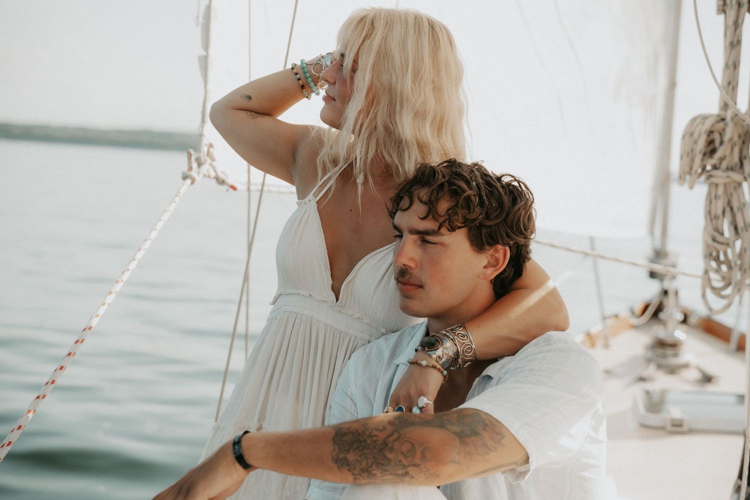 Couple embracing on a sailboat, woman in white dress with blonde hair, man with tattoos, ocean background.