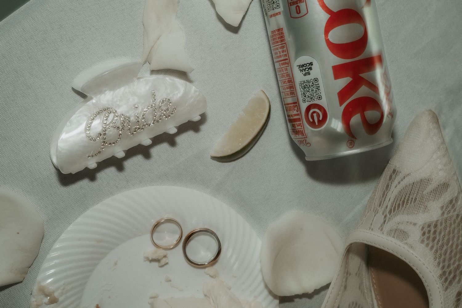 Wedding rings on a plate beside a Diet Coke can, seashells, and lime slices on a white surface.