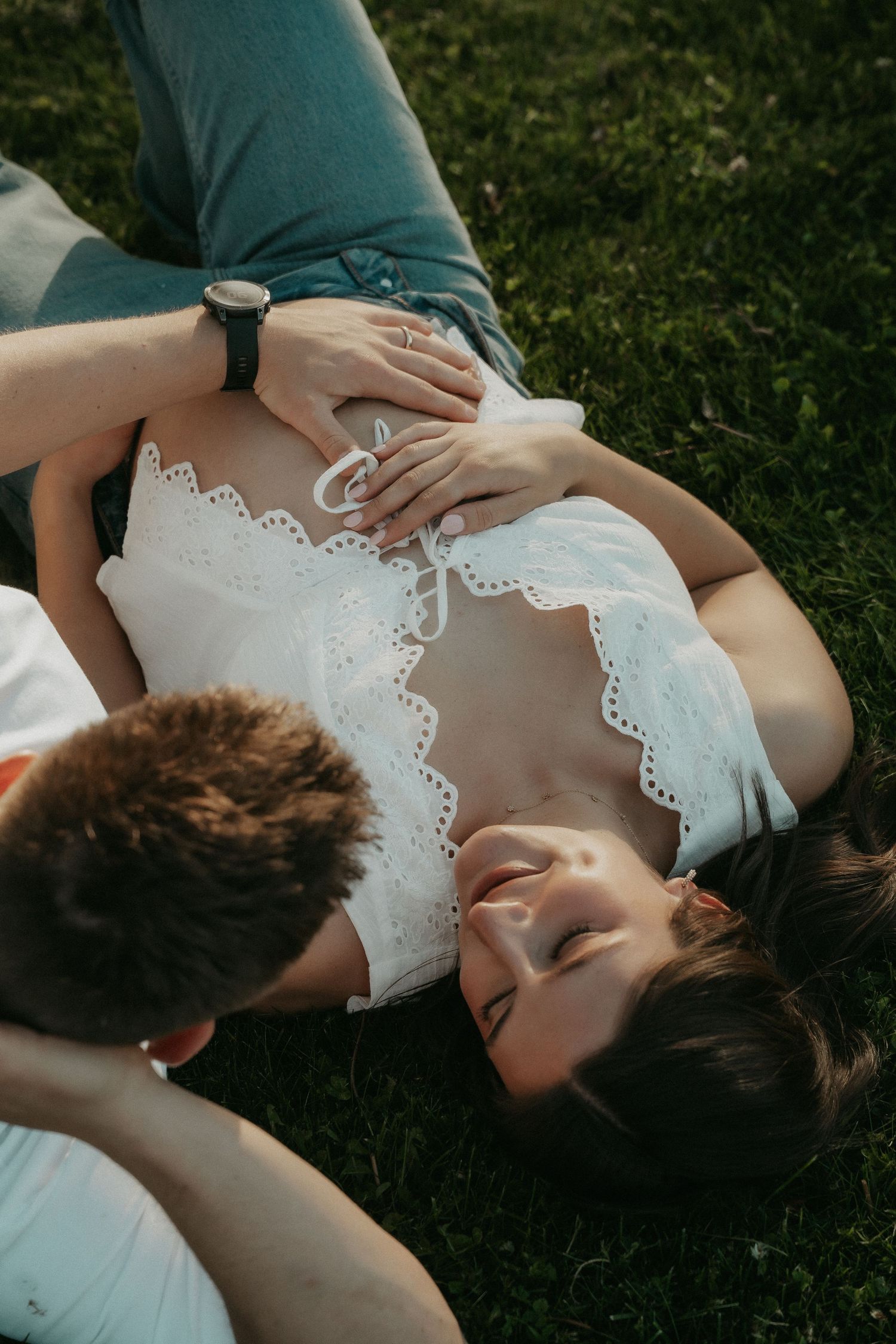 Couple lying on grass, woman in white top smiling, partner placing hands on her pregnant belly outdoors.
