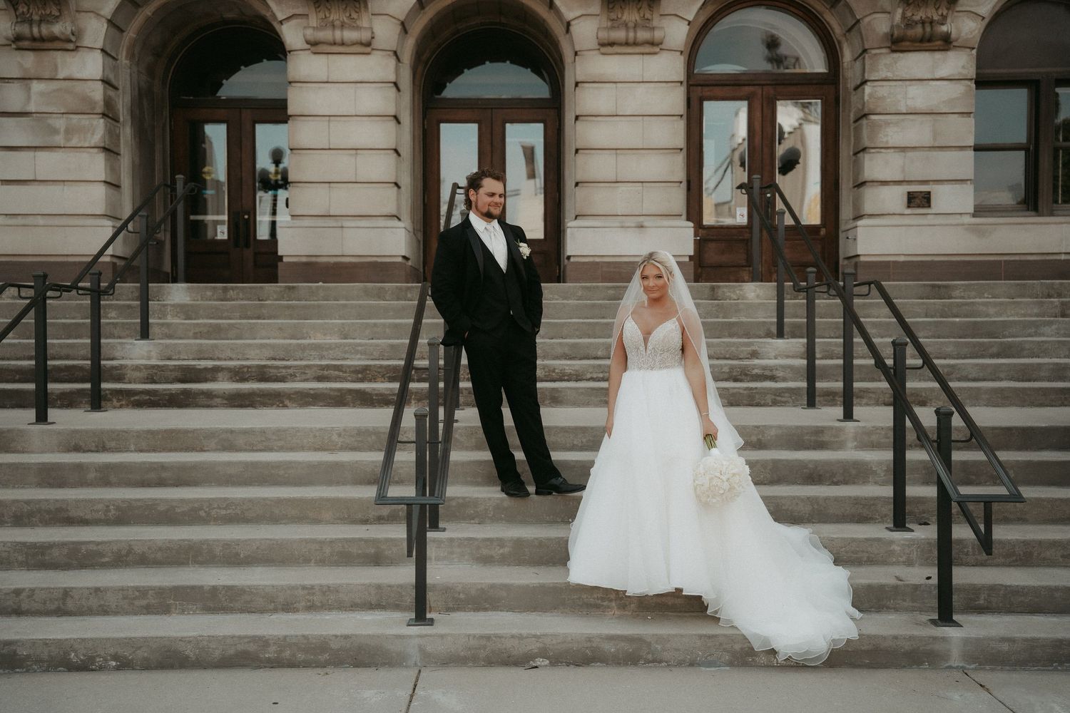 Elegant bride and groom pose on stone steps outside historic building during wedding photoshoot.