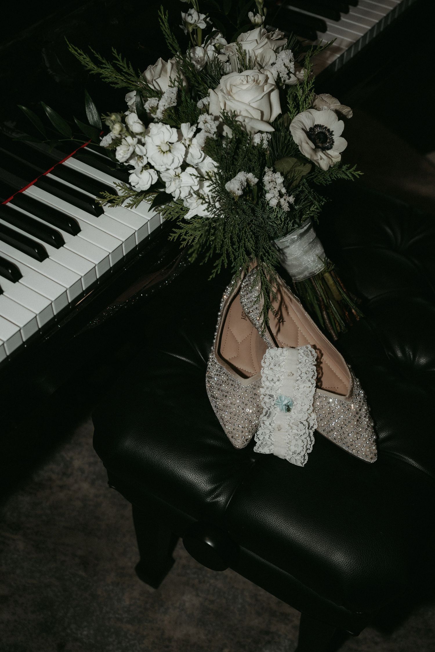 White bridal bouquet with roses and anemones rests on piano keys next to sparkly wedding shoes.
