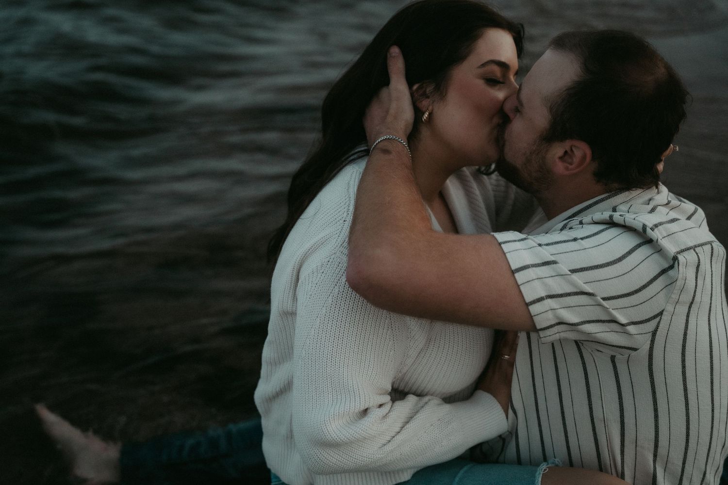 A romantic couple sharing an intimate kiss by dark waters at dusk.
