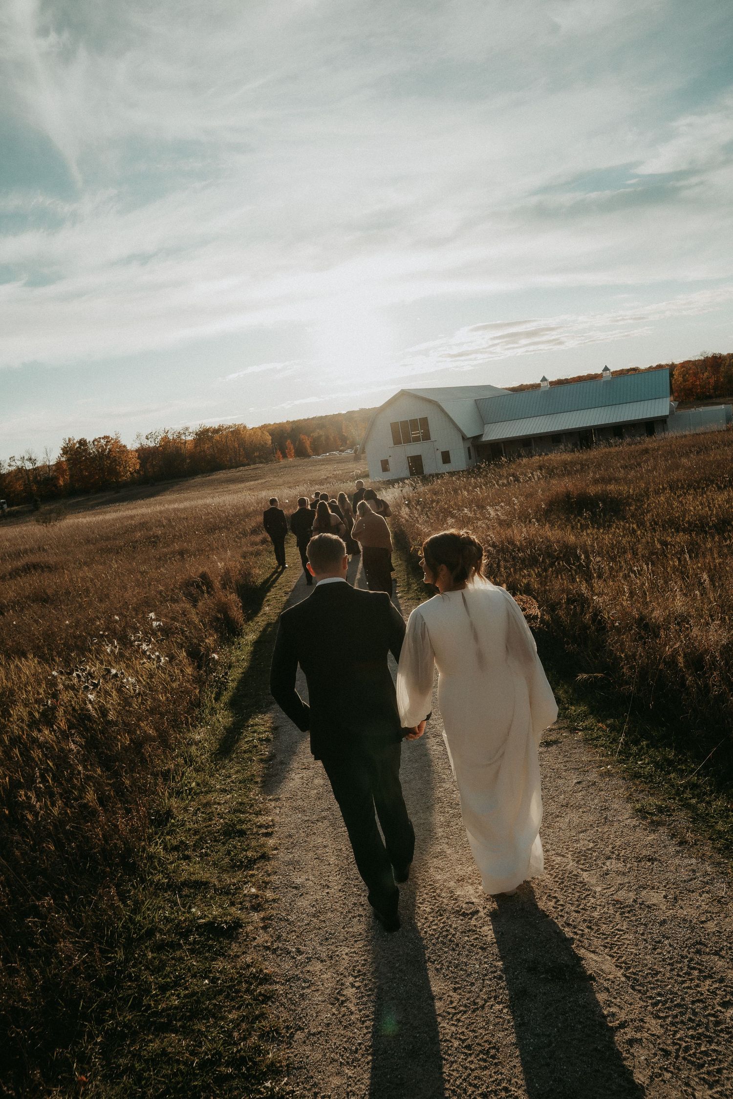 A couple walks down a dirt path towards a white barn at sunset, casting long shadows in the golden light.