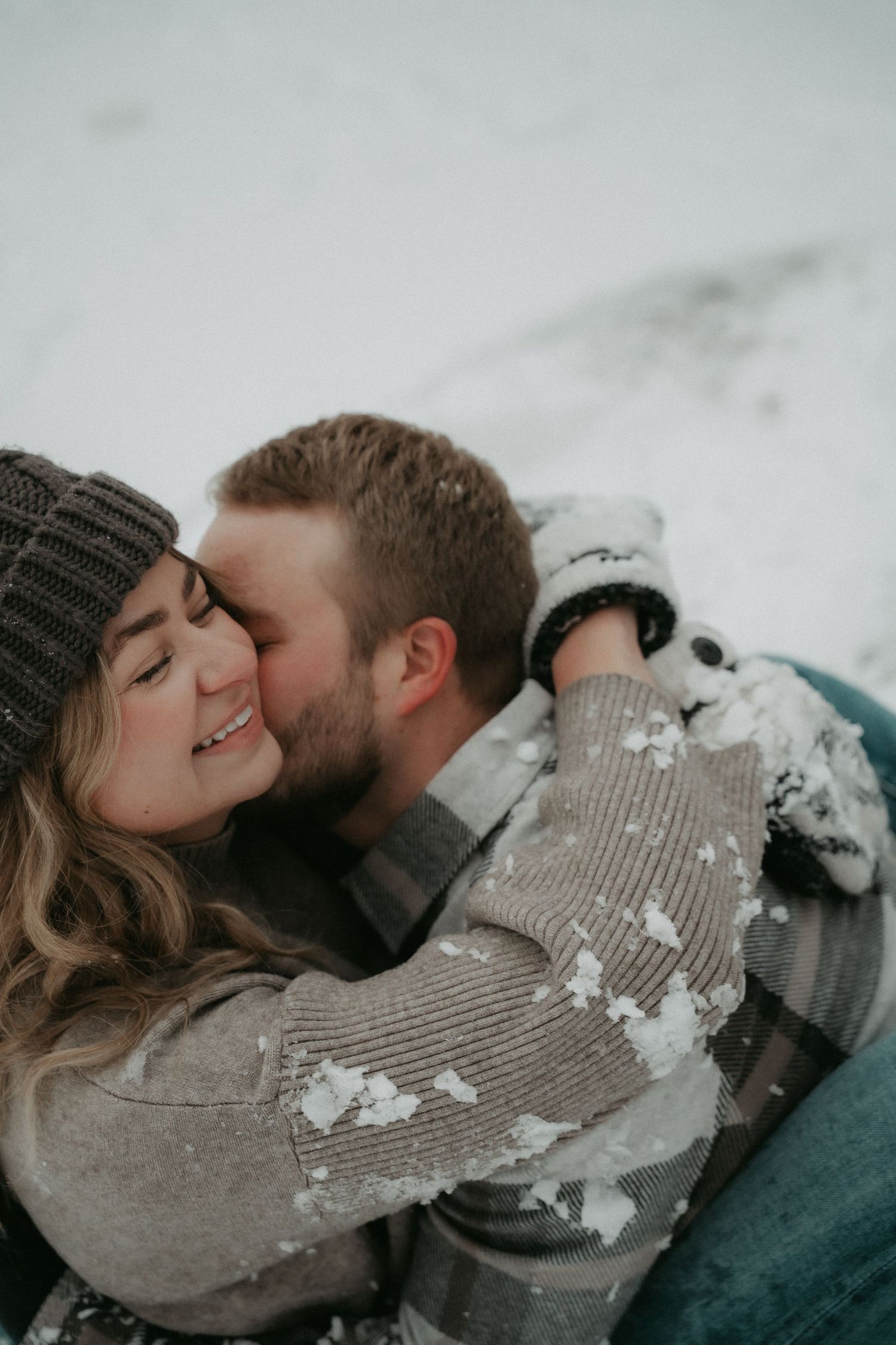 A romantic winter scene shows a couple laying in the snow sharing intimate moments in cozy sweaters.