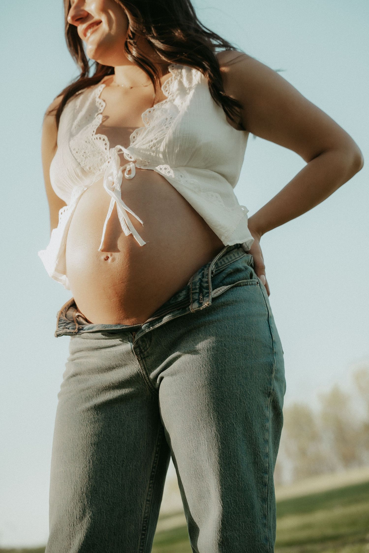 Pregnant woman in white crop top and jeans showing baby bump outdoors in natural sunlight.