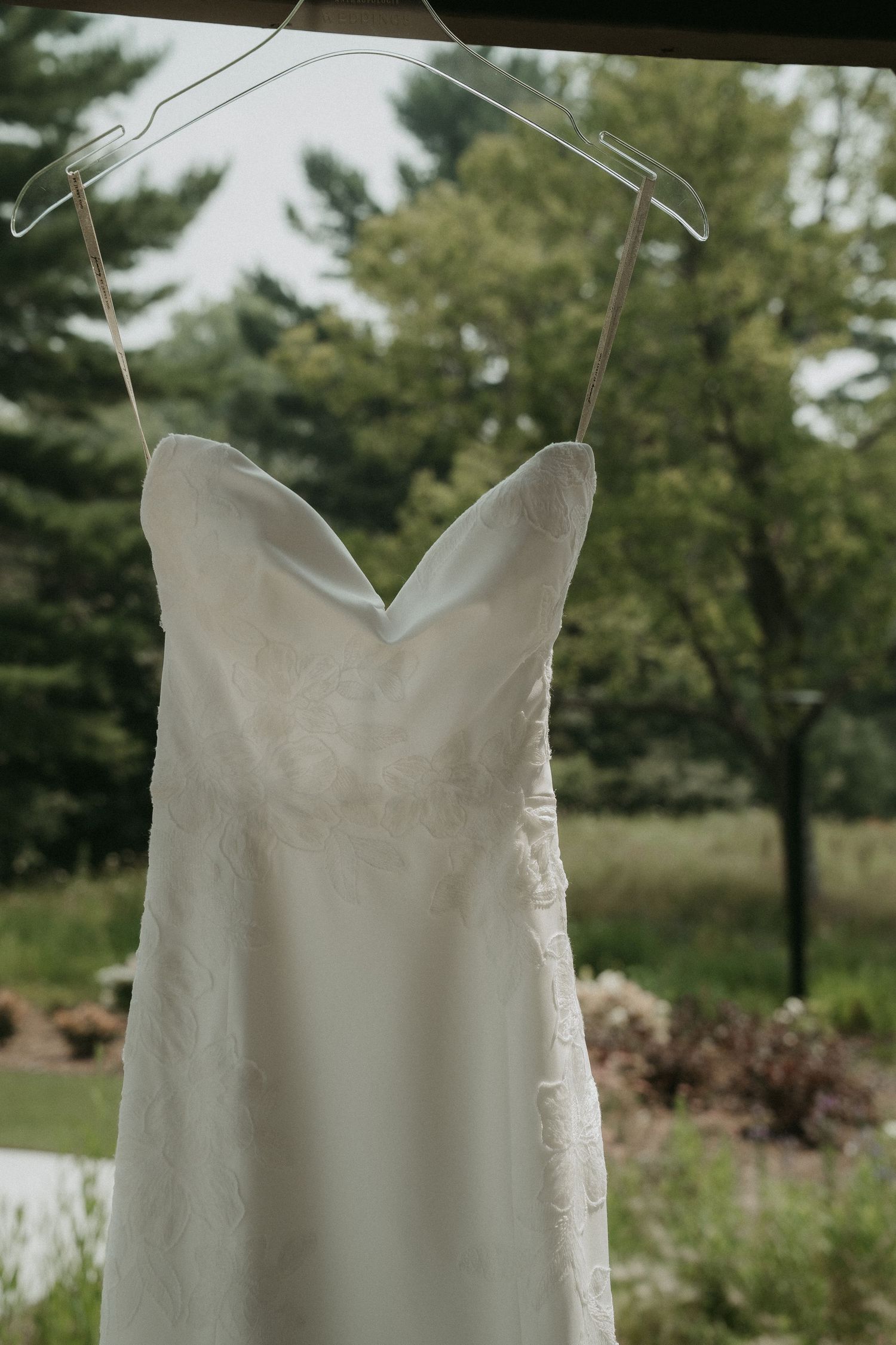 White strapless lace wedding dress hanging on a clear hanger against a lush green outdoor backdrop.