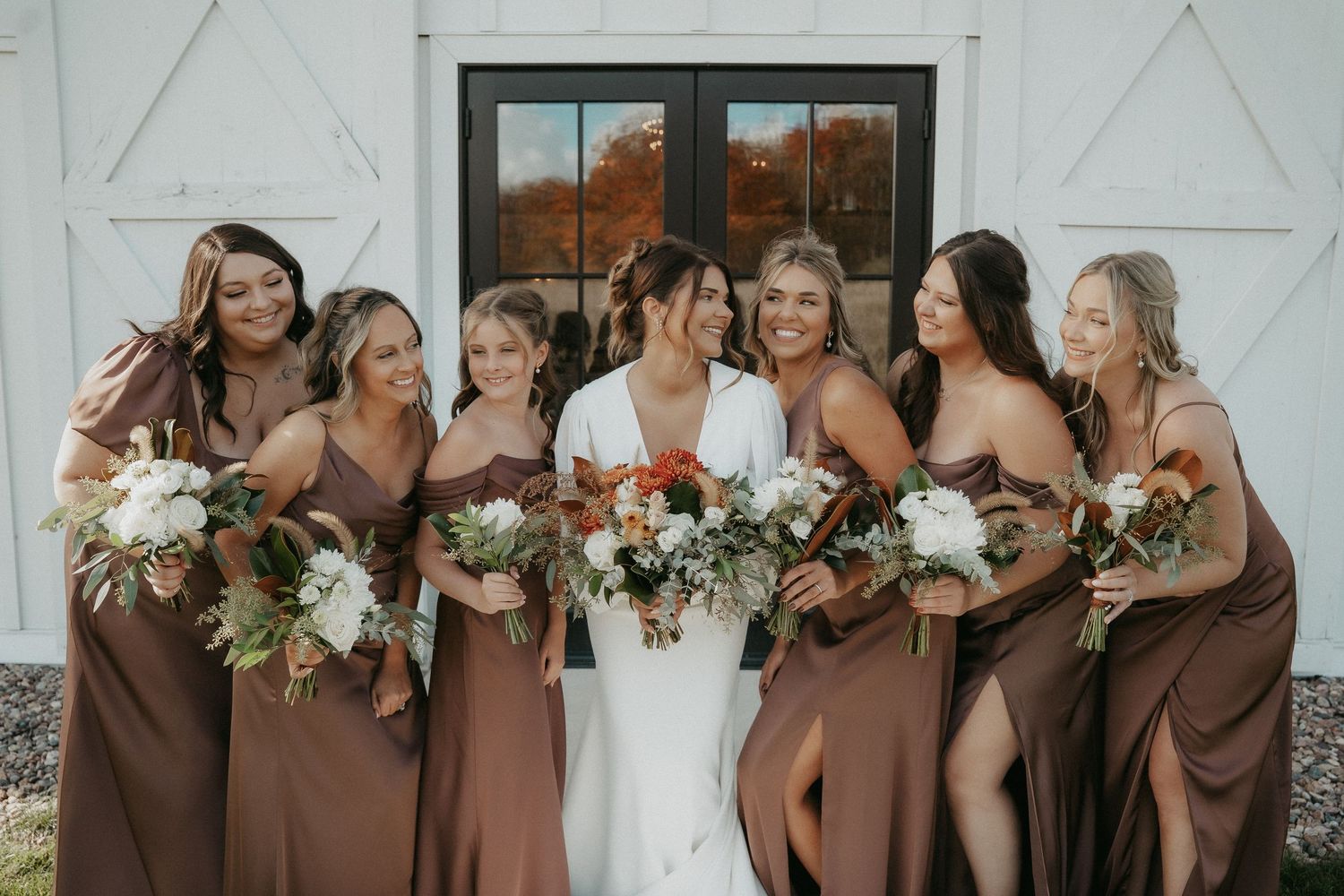 A group in brown bridesmaid dresses and white wedding gown pose together at a rustic barn venue.