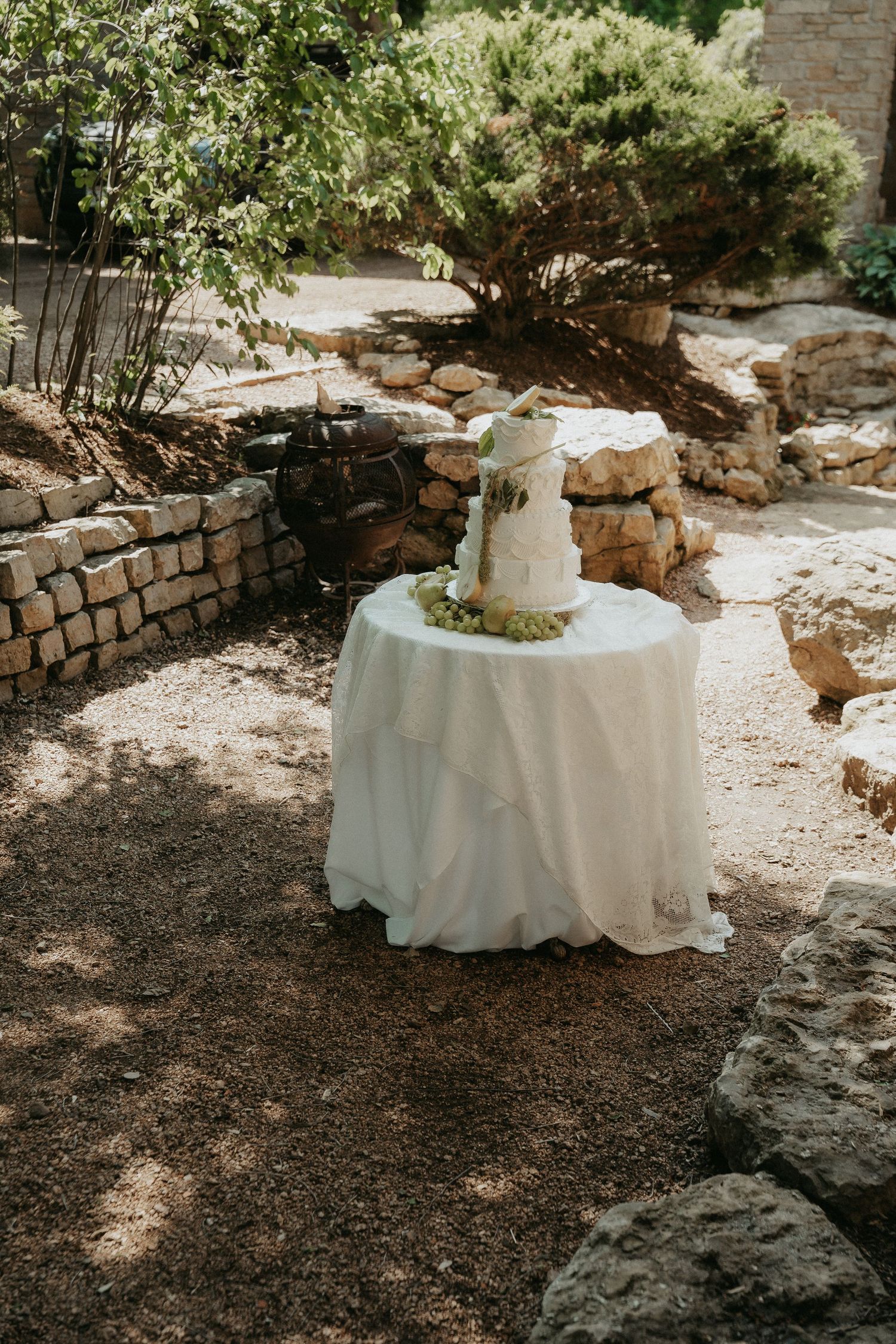 White tiered wedding cake on draped table in rustic outdoor garden with stone walls and lush greenery.