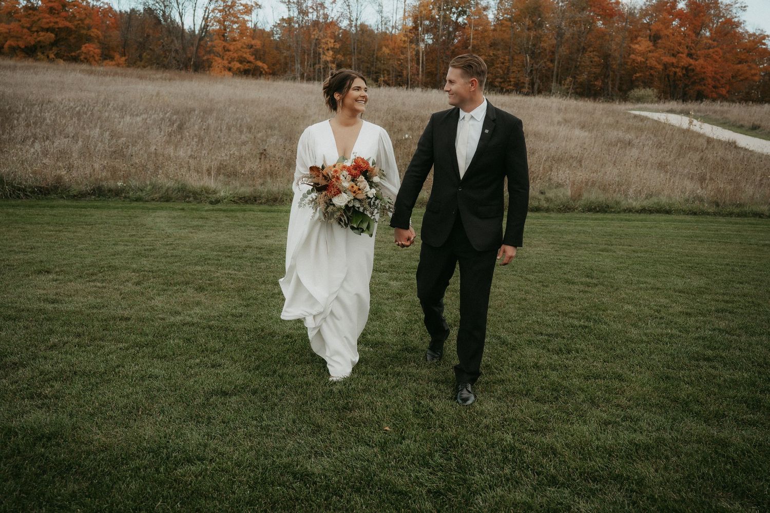 A couple walks hand in hand through an autumn field during a wedding celebration as fall foliage glows in the background.