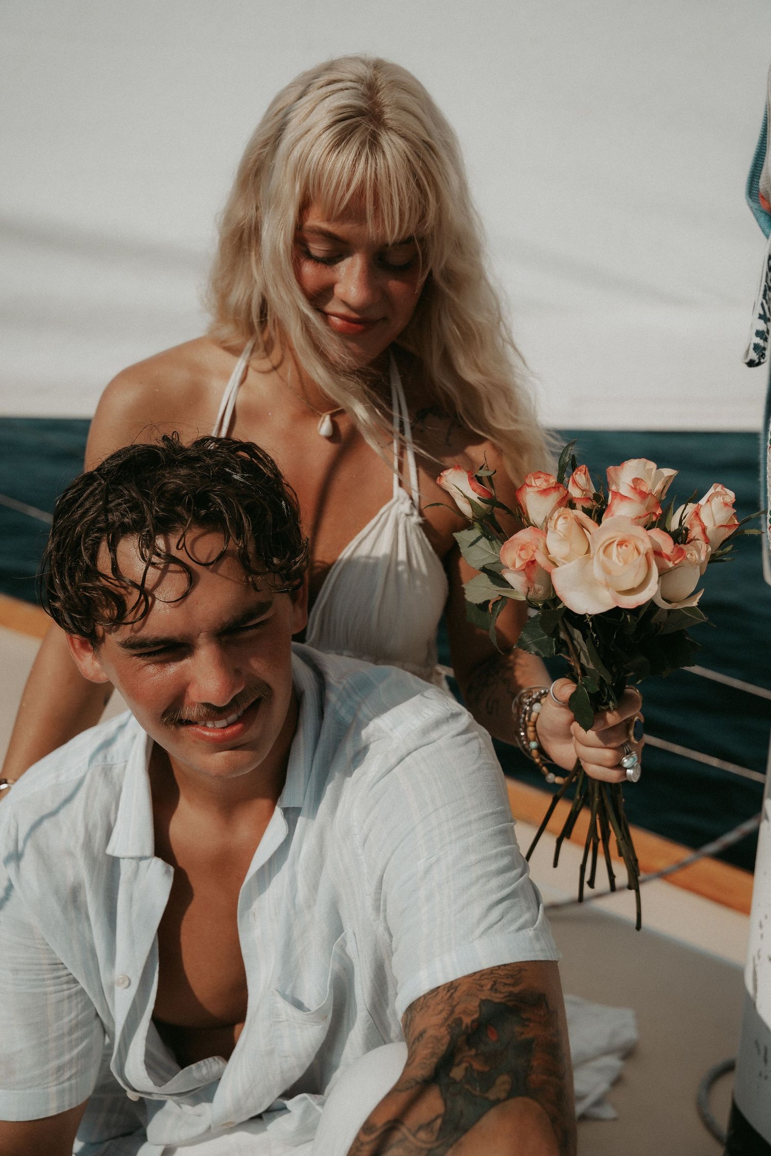 Couple on a yacht, woman in white bikini holding pink roses bouquet, man in white shirt with tattoo smiling.