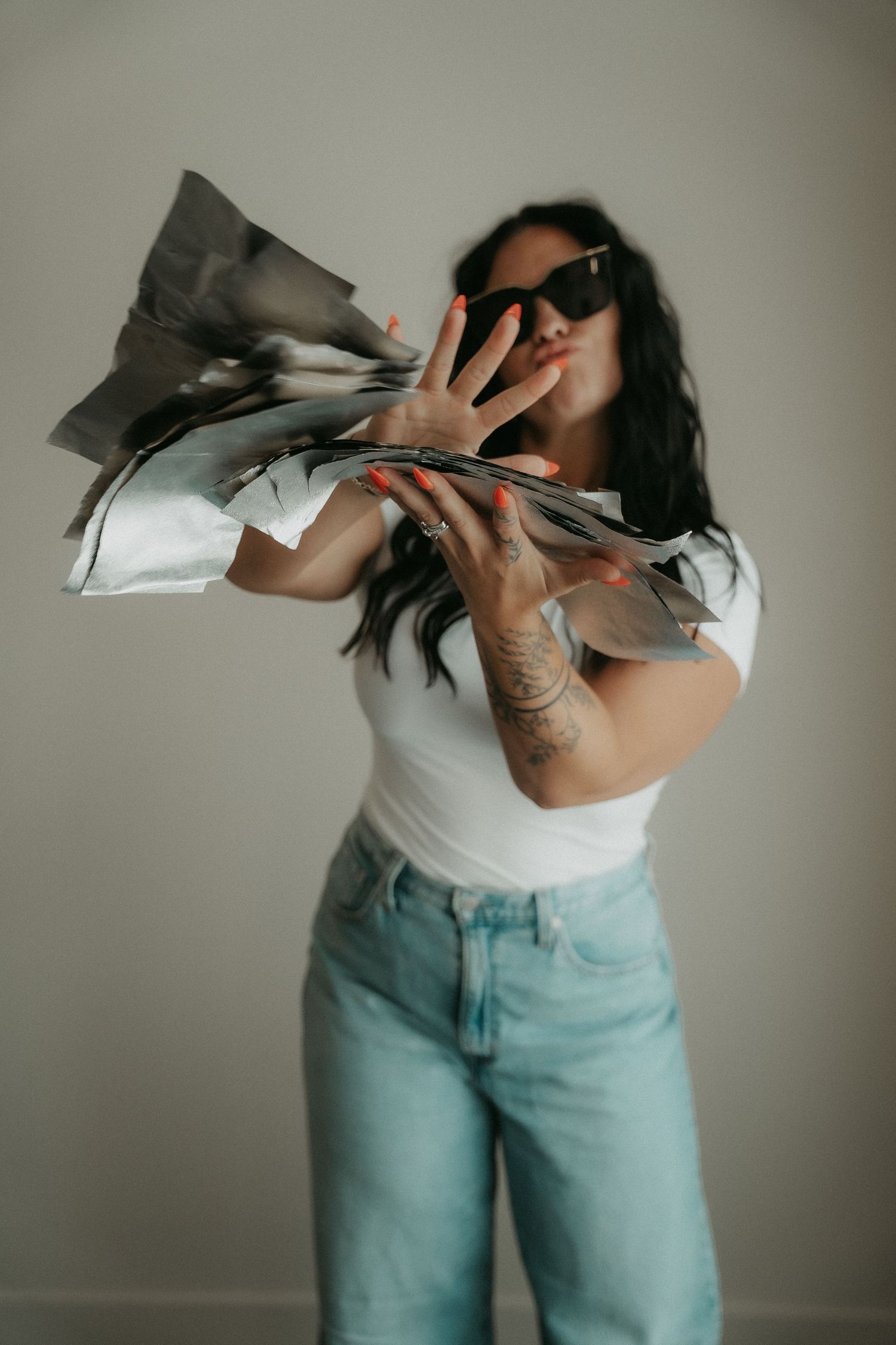 Woman in white tee and jeans tossing papers, wearing sunglasses with a hand tattoo visible.