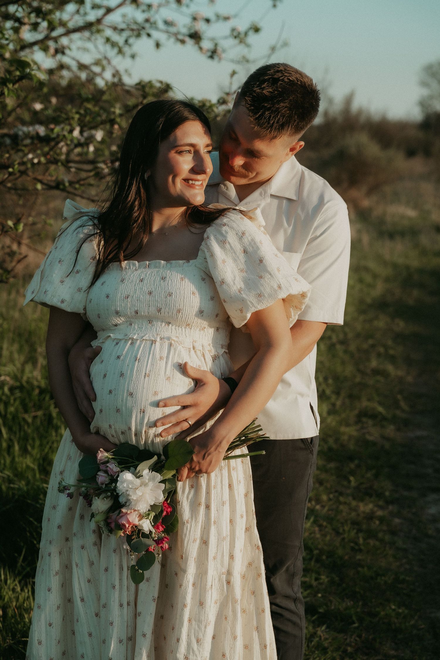 Couple sharing a tender maternity moment outdoors, woman in floral dress holding baby bump with bouquet in golden sunlight.