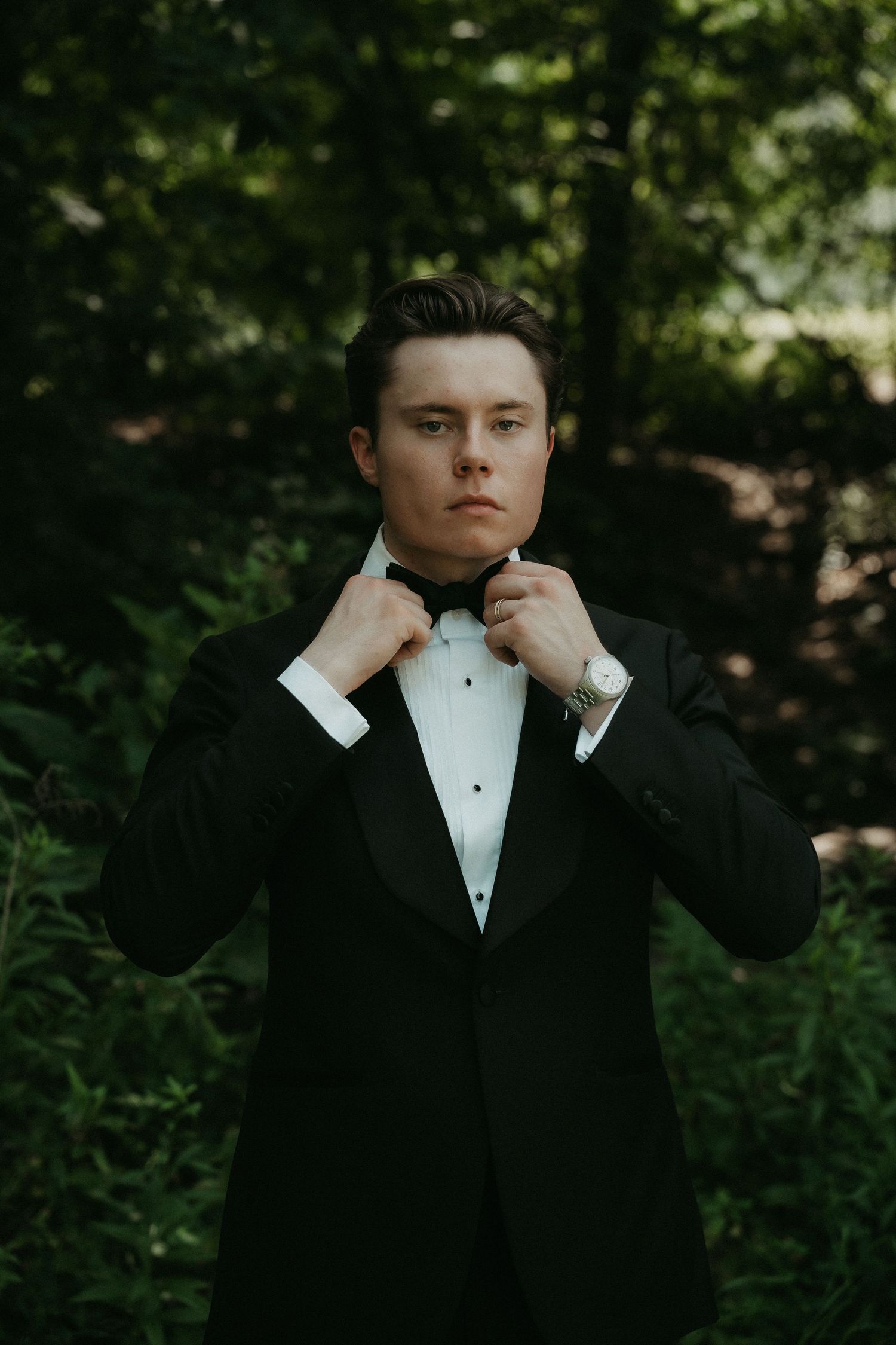 Young man in black tuxedo adjusting bow tie outdoors with lush green forest background, formal wedding attire.
