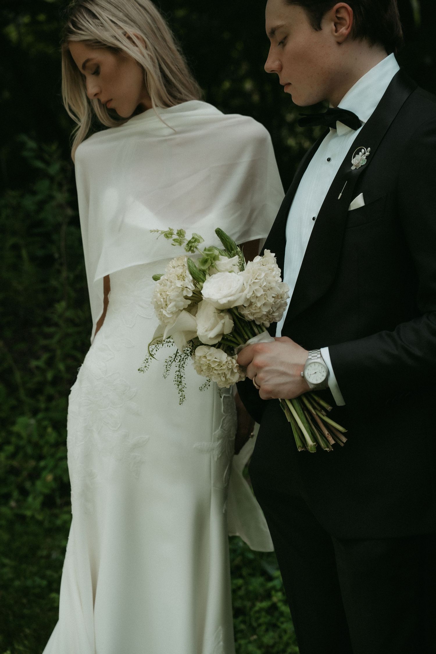 Bride in white cape gown holds bouquet with groom in black tuxedo, posing before lush green foliage.