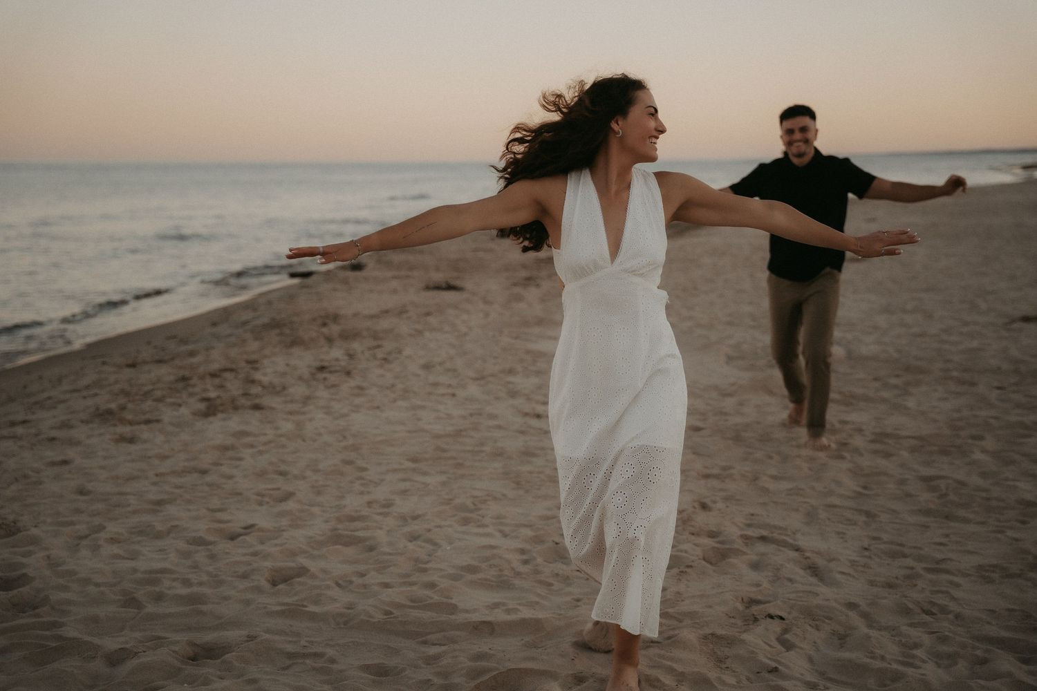 A couple walks hand in hand along a sandy beach at sunset wearing white and brown casual attire.
