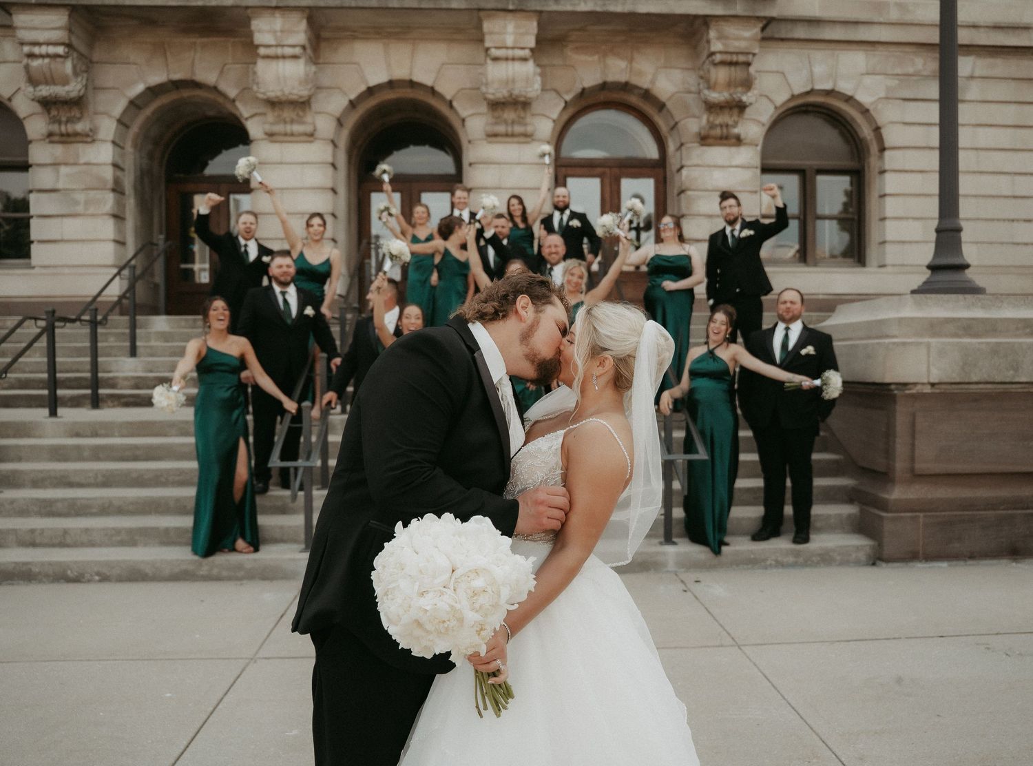 Wedding party members pose and celebrate together on grand stone steps of historic building.