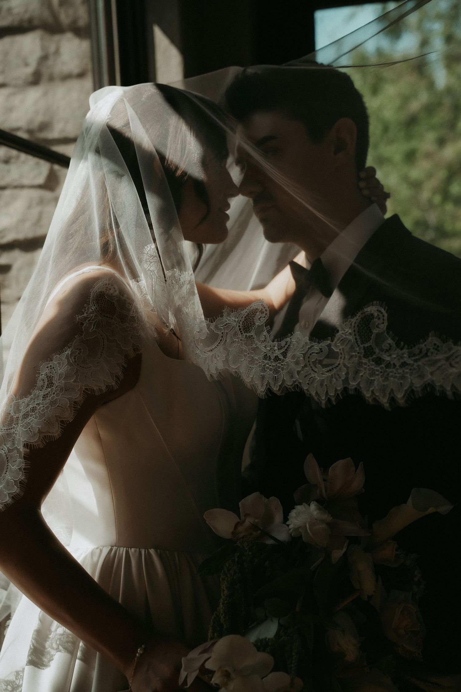 Bride and groom share an intimate moment under a lace veil, bride holding a floral bouquet near a stone wall.