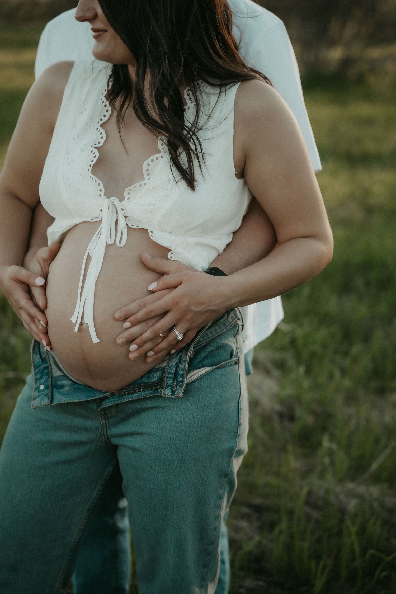 Pregnant woman in white crop top and jeans cradling baby bump outdoors in a maternity photo session.