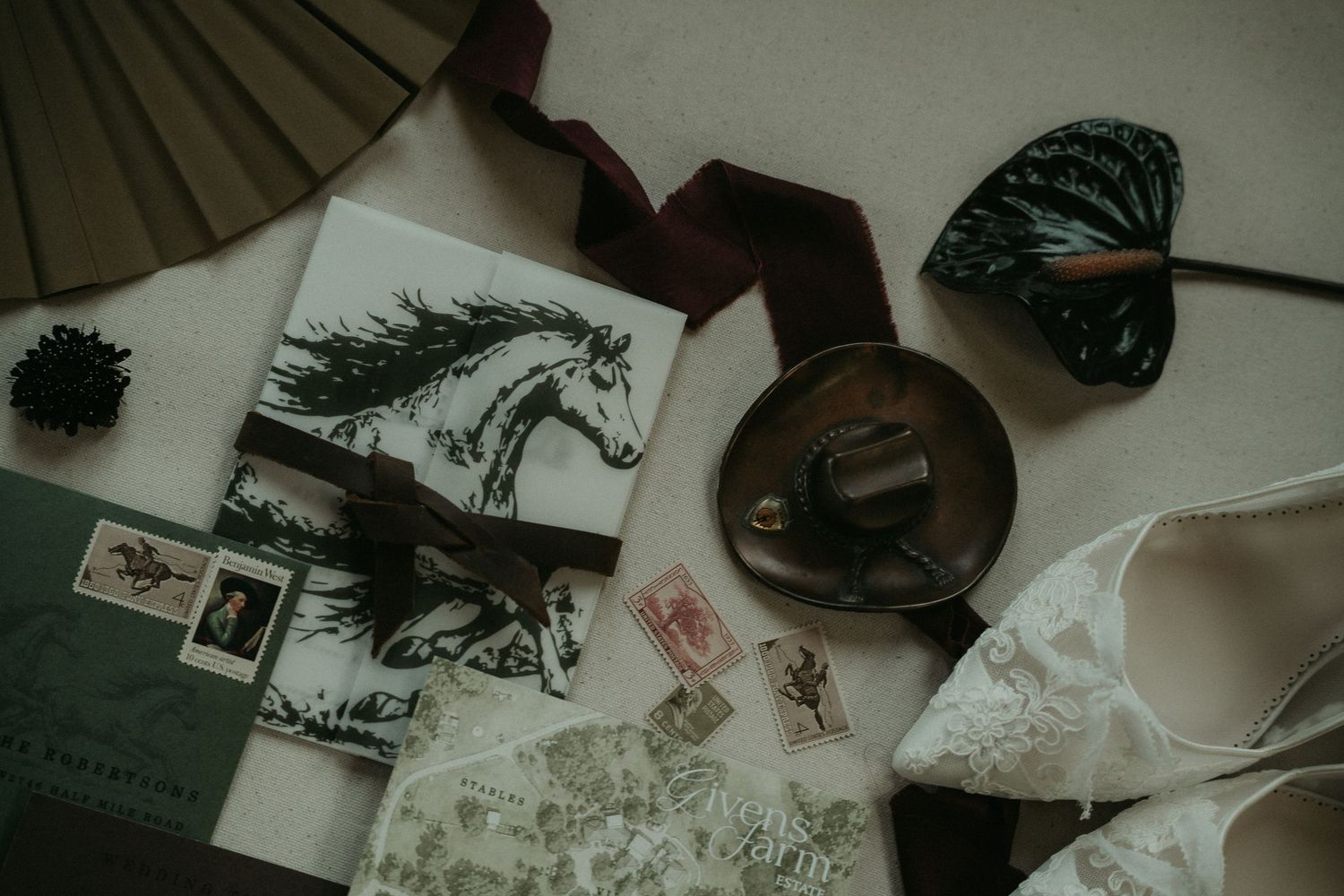 Flat lay of wedding stationery with horse illustration, vintage stamps, fan, ribbon, and lace bridal shoes on neutral background.