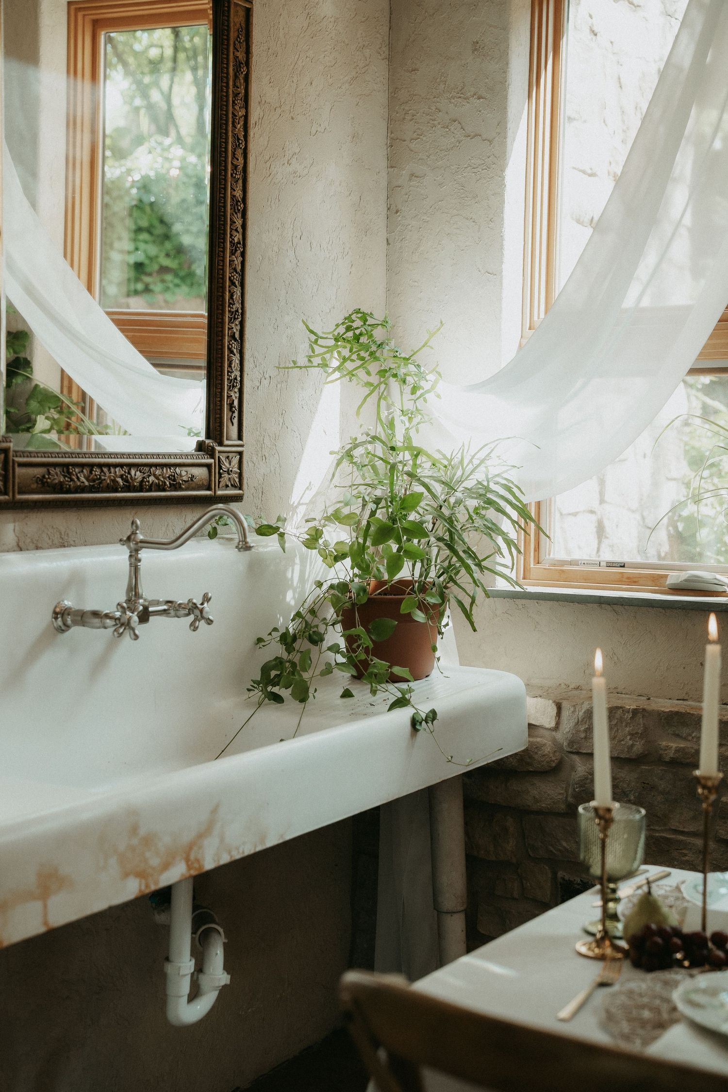 Vintage bathroom with ornate mirror, white sink, potted green plant, lit candles, and sheer curtains by a window.