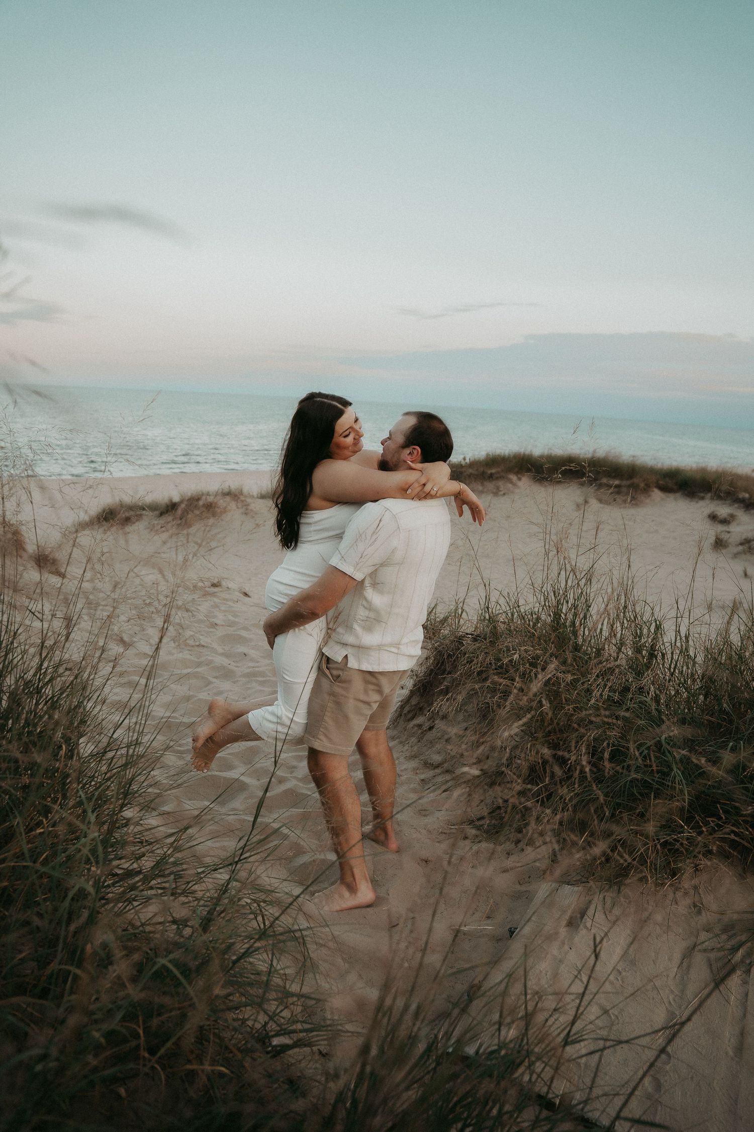 A couple in white outfits dances together on a sandy beach dune at sunset, creating a romantic silhouette.