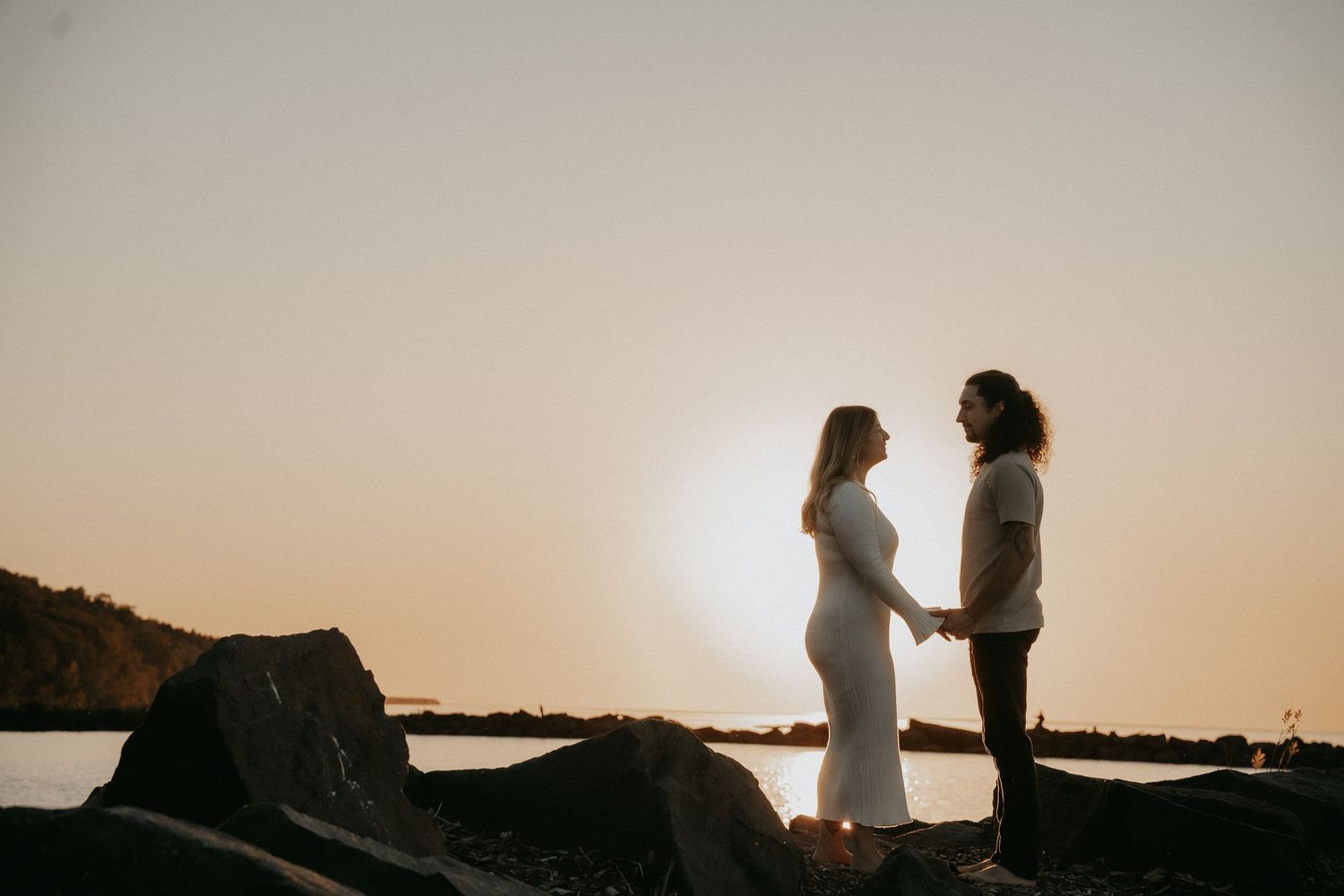 A silhouetted couple stands on rocky shore at sunset with ocean and mountains in background.