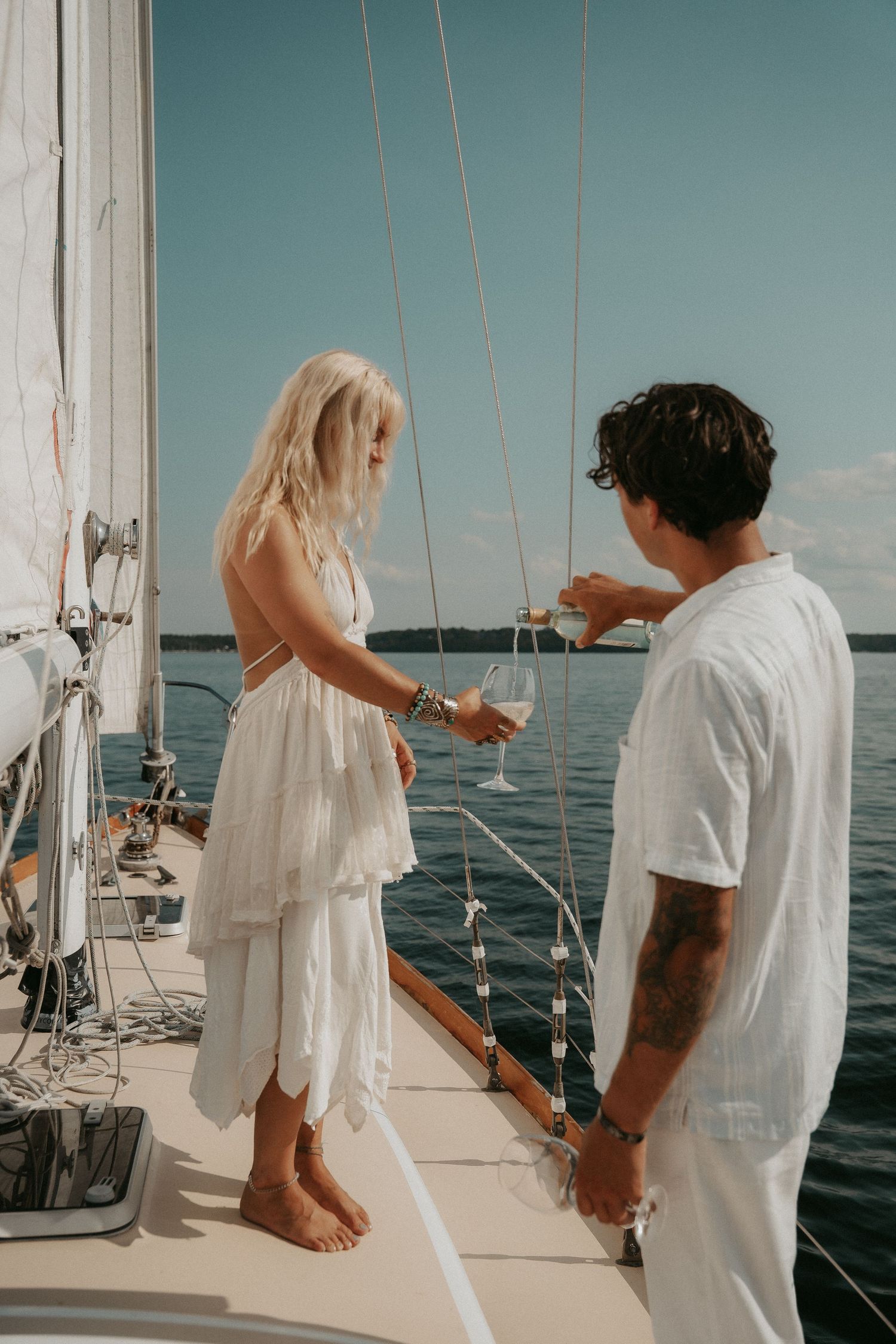 Couple in white outfits toasting with wine glasses on a sailboat deck with calm blue ocean in the background.