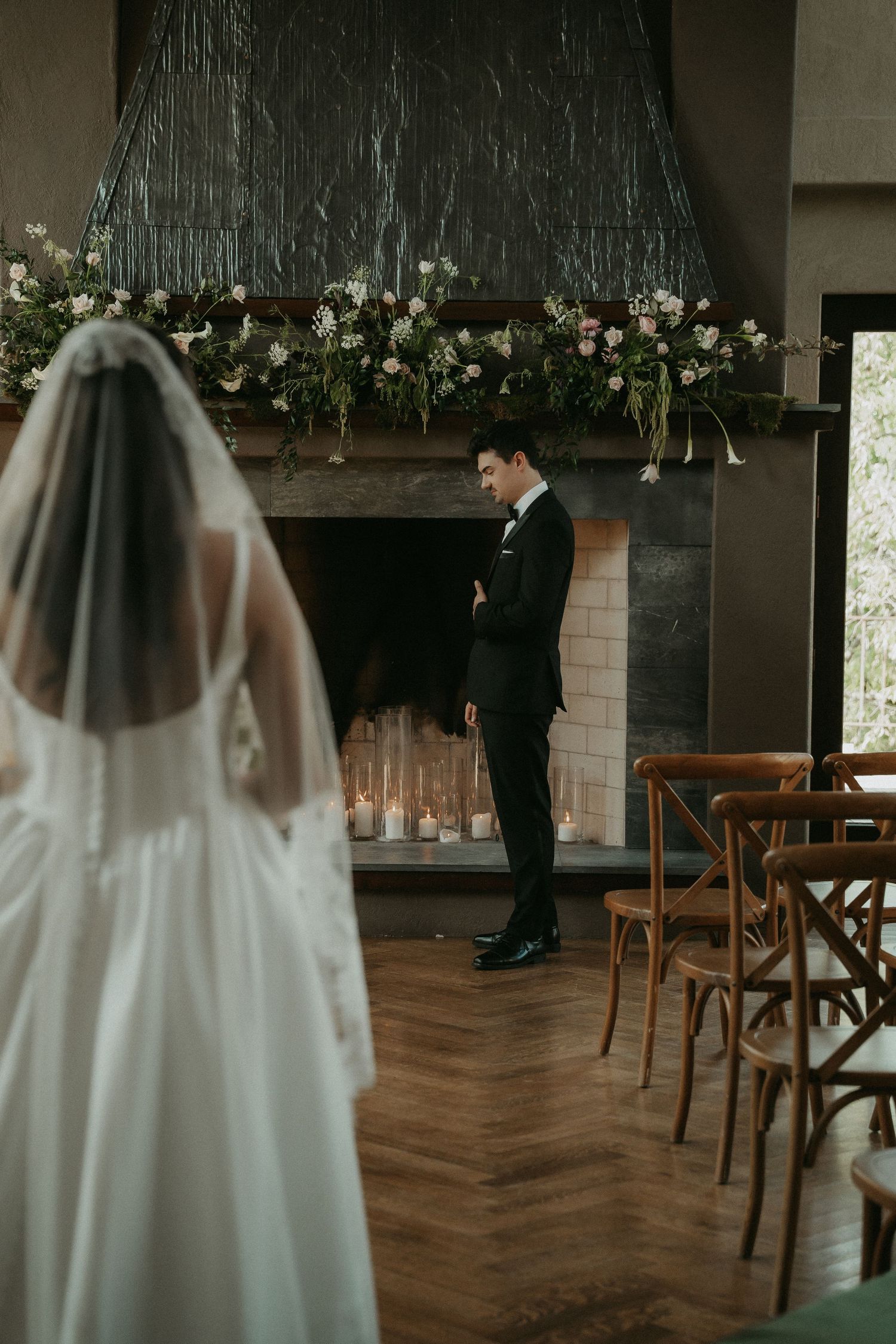 Groom in black tuxedo stands at floral fireplace altar as bride in white gown and veil approaches during indoor wedding ceremony.