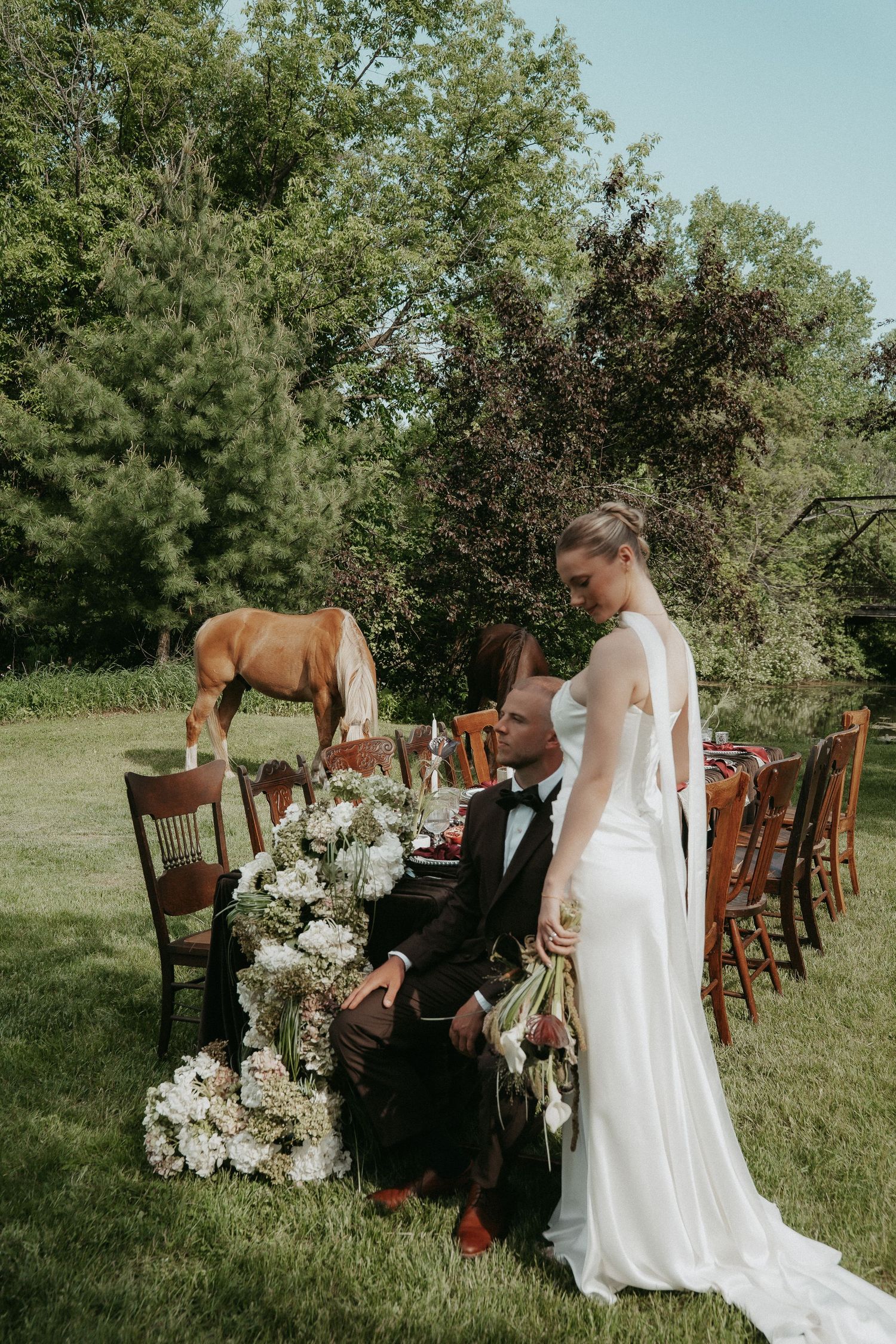Bride in white gown stands beside groom at outdoor wedding table with floral arrangements and a horse in the background.