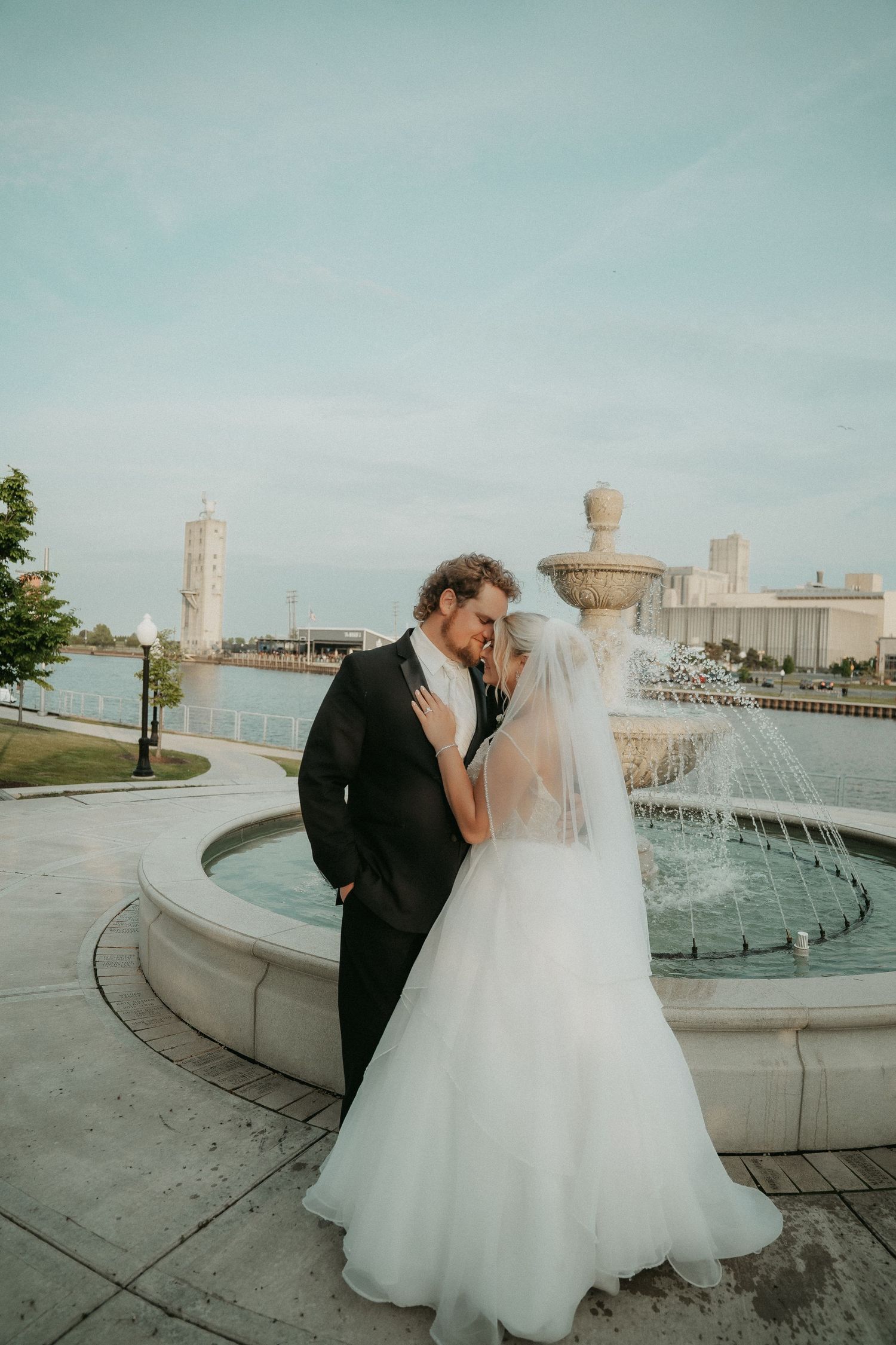 A couple shares a romantic moment by a curved concrete wall overlooking a waterfront on their wedding day.
