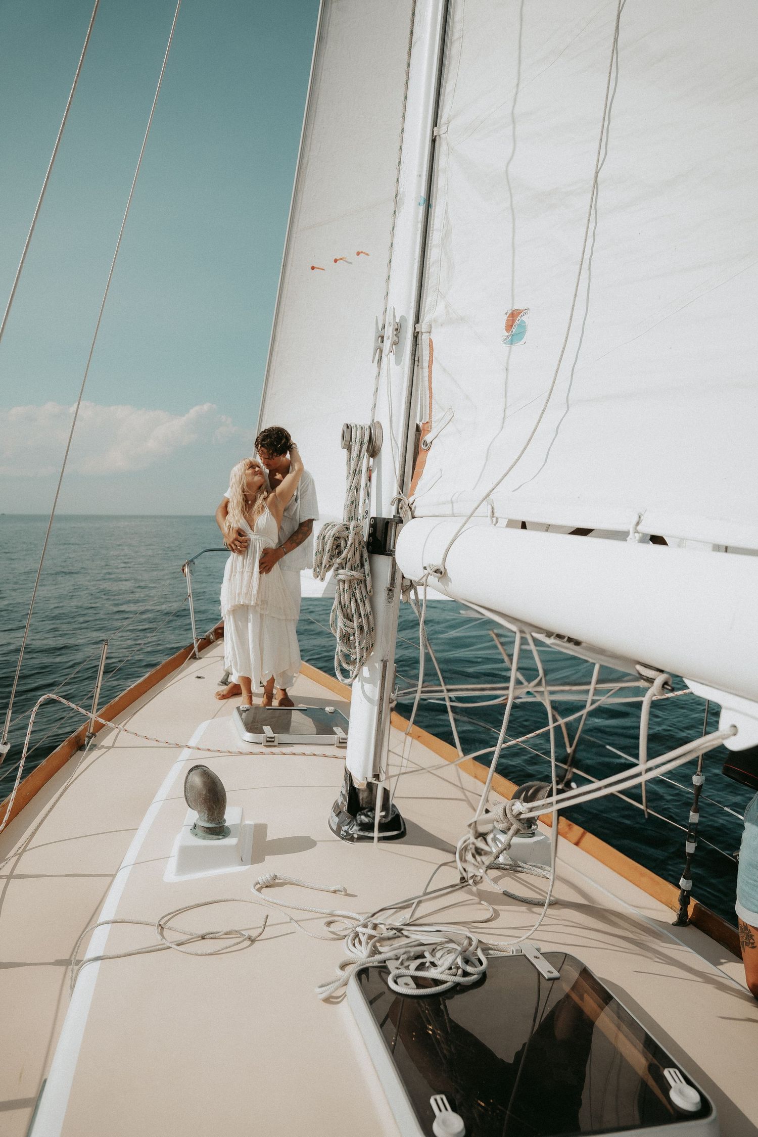 Couple embracing on the deck of a white sailboat on calm blue ocean waters on a sunny day.