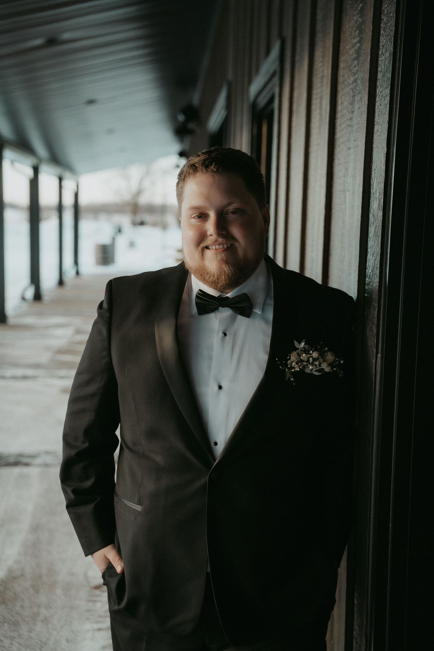A groom in a classic black tuxedo and bowtie poses in a dimly lit hallway with one hand in his pocket.