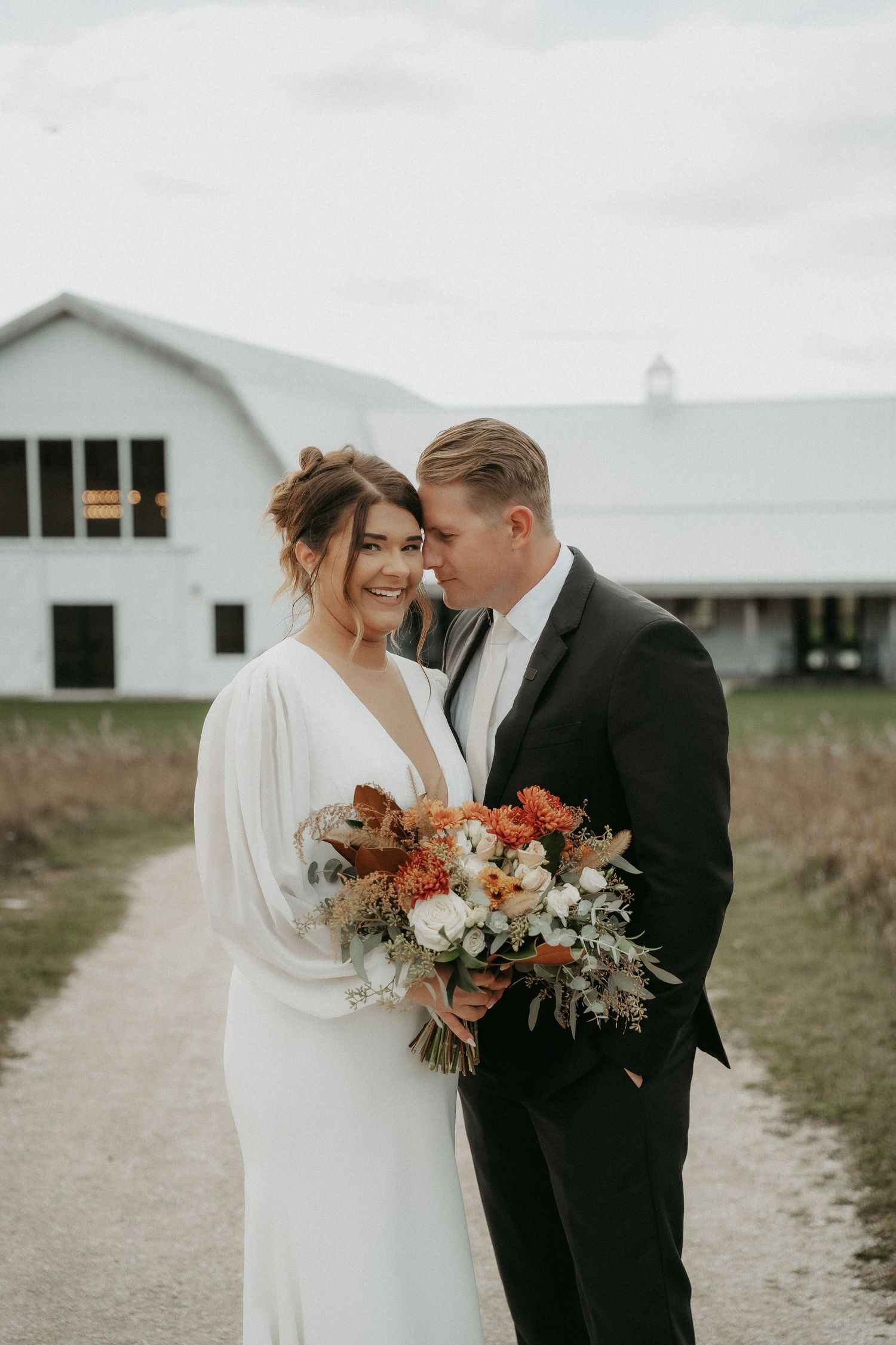 A couple shares an intimate moment on their wedding day holding a beautiful autumn-toned bouquet near a rustic barn.
