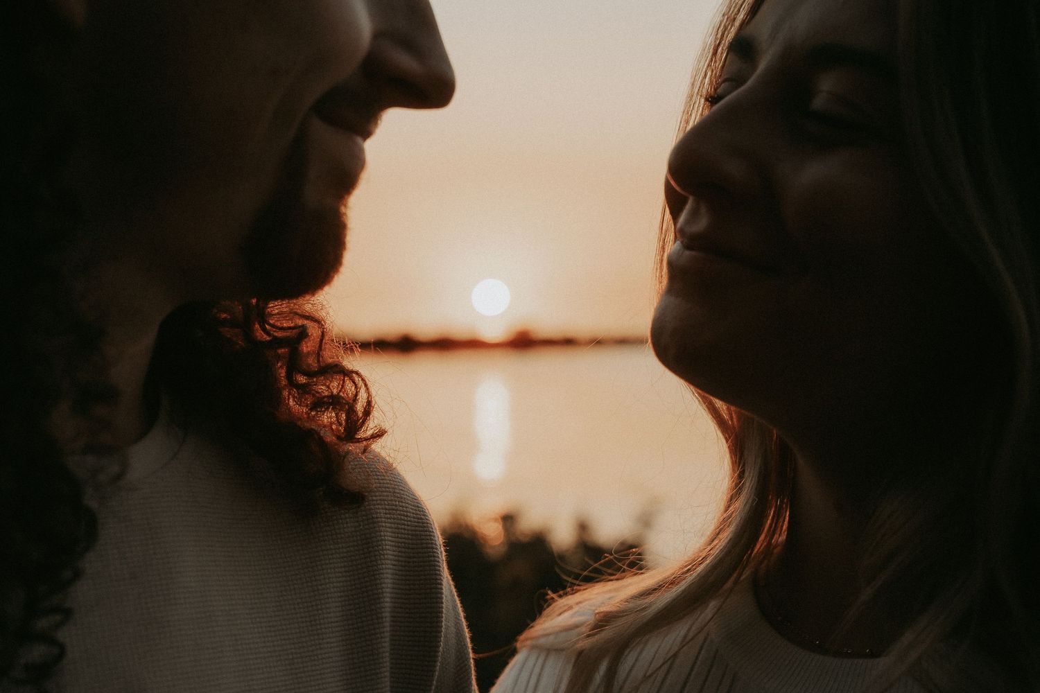 Silhouetted couple embraces against a glowing orange sunset over a calm lake.