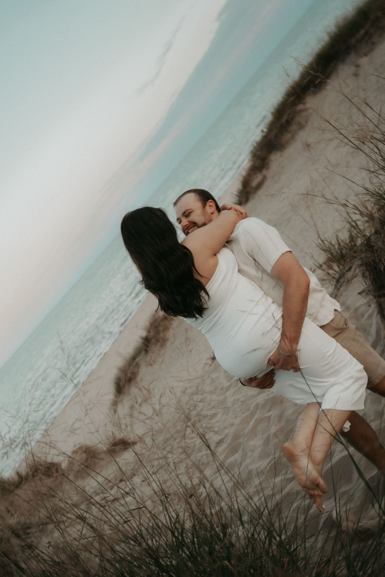 A romantic couple in white outfits embrace on a sandy beach dune with ocean waves and cloudy sky in the background.