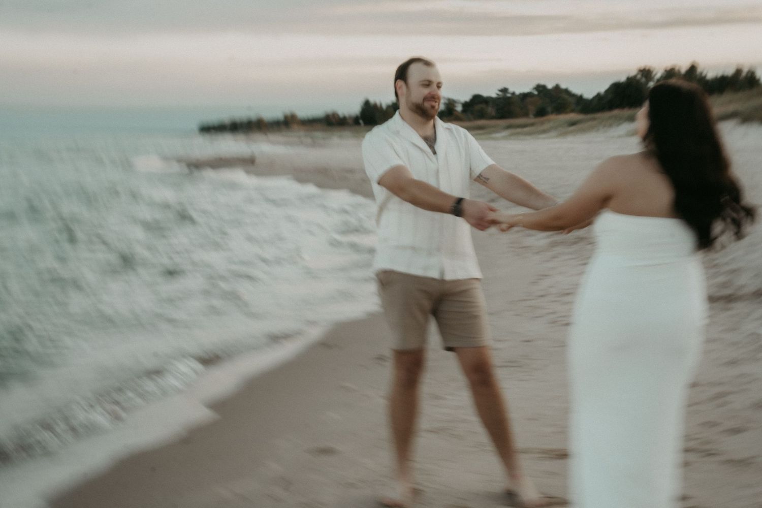 A couple in light colored outfits dances and plays together on a sandy beach at sunset.