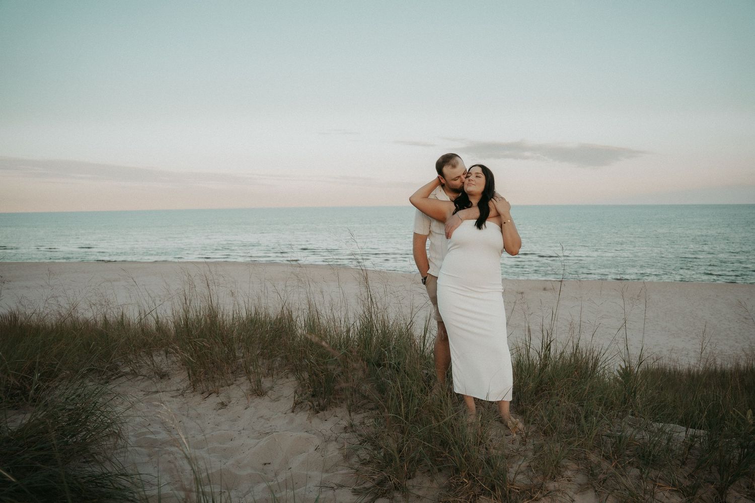 A couple embraces on a sandy beach with tall grass and ocean waves in the background at sunset.