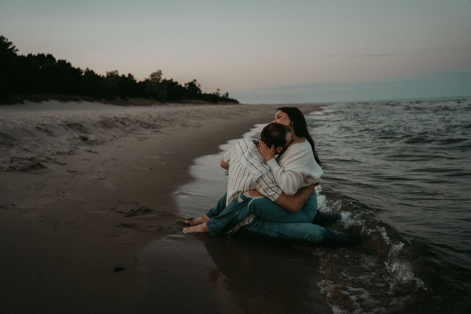 A person sits on a sandy beach at sunset wearing jeans and a white shirt beside the ocean waves.