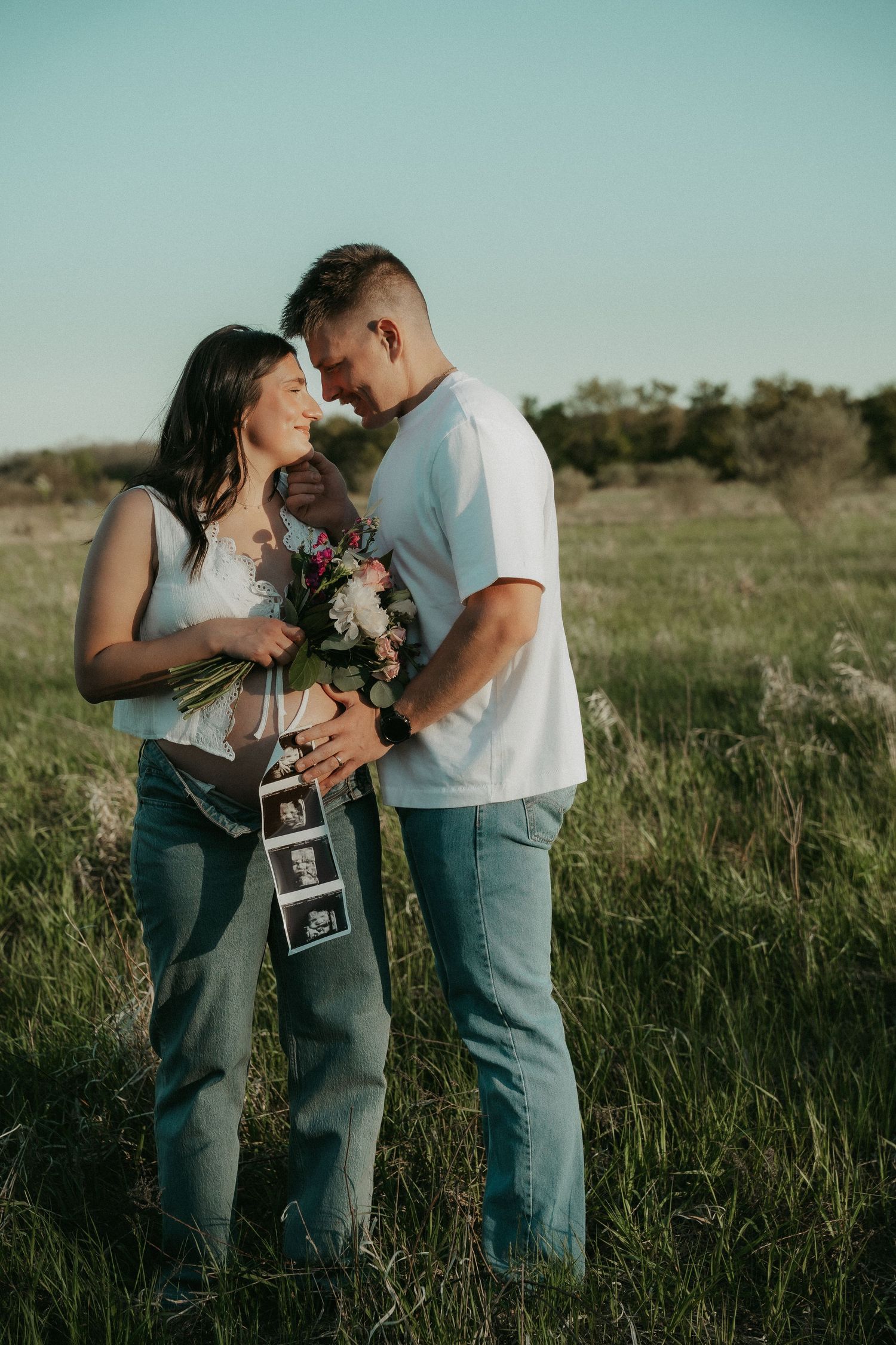 Couple sharing a tender moment in a field, holding ultrasound photos and flowers, announcing their pregnancy.