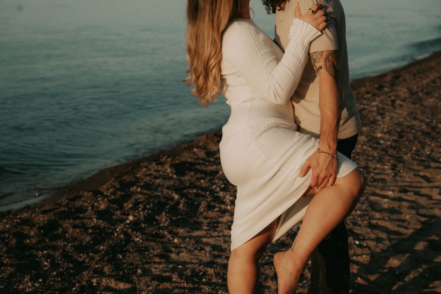 A woman in a white dress poses on a rocky beach at sunset with water in the background.